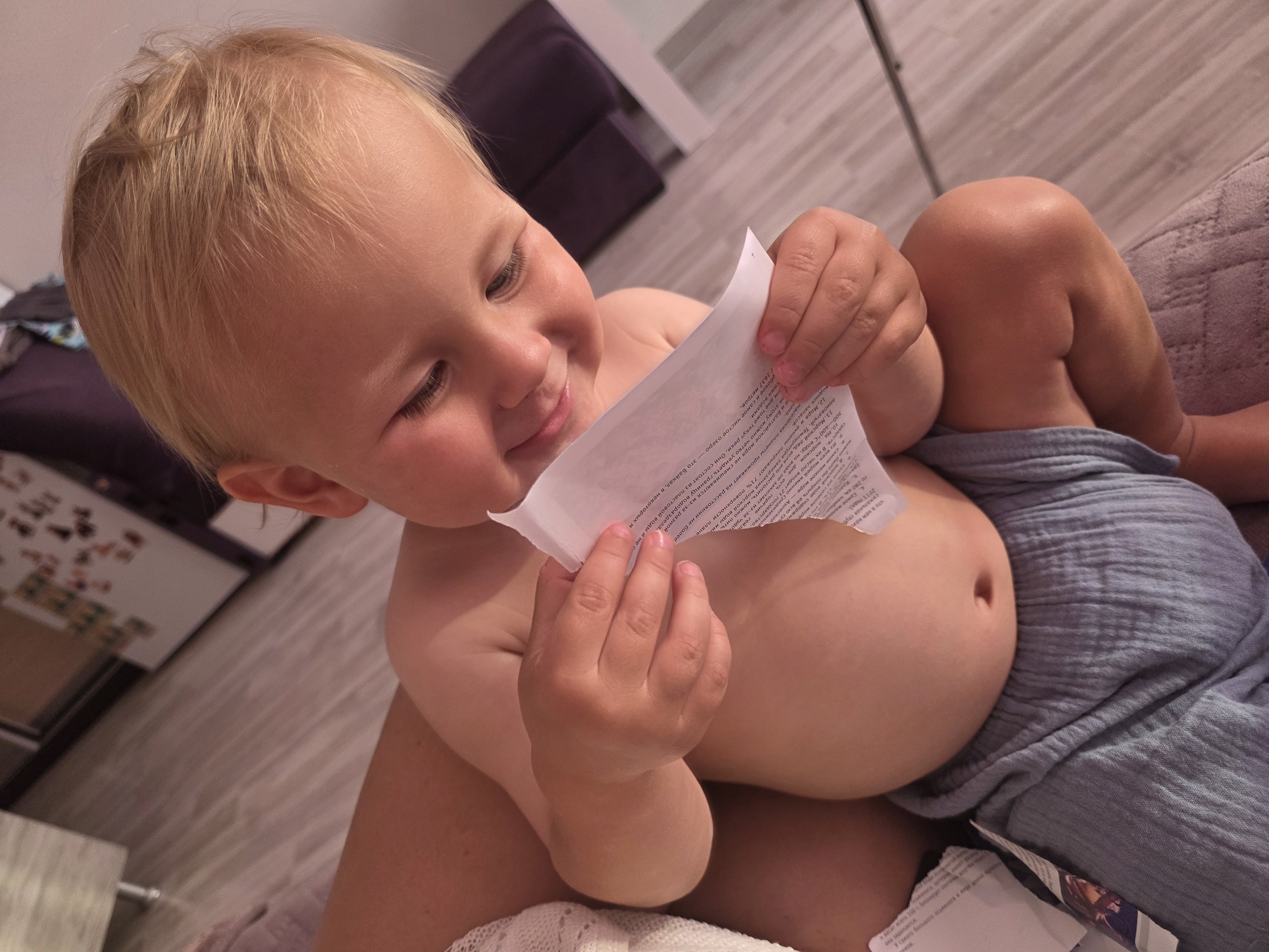 A close-up shot shows a young blond child sitting on a person's lap, holding a piece of paper with text on it. The child is looking down at the paper with a happy and engaged expression, their eyes crinkling at the corners. The child's skin is rosy, and they are wearing a light blue diaper or cloth wrap around their lower body. The background is softly blurred, showing what appears to be a wooden floor and furniture, suggesting an indoor setting. The lighting indicates it might be evening or a dimly lit room. The text on the paper appears to be in Russian, with some visible numbers and words. The overall mood is one of a quiet, intimate moment between the child and the person holding them, as the child explores the paper.