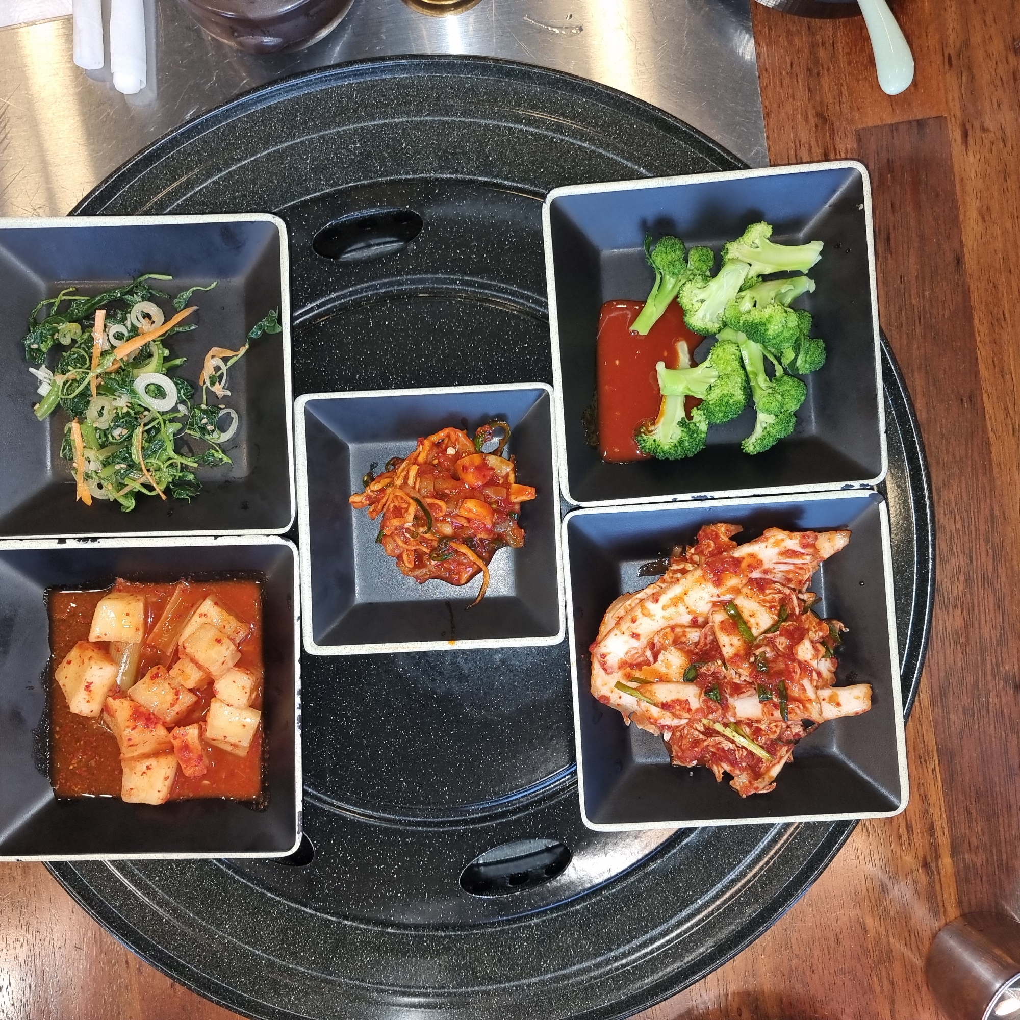 In Dunnae-myeon, South Korea, a top-down shot displays an assortment of Korean side dishes (banchan) arranged on a black, circular grill mat. The dishes are presented in square, black bowls with off-white rims. The top left bowl contains diced white radish kimchi (kkakdugi) in a vibrant red sauce. To its right, the top right bowl holds a salad of mixed greens, likely seasoned with sesame oil and chili flakes, featuring julienned carrots and white onions. In the center, a smaller square bowl contains a portion of what appears to be stir-fried squid or other seafood with vegetables in a reddish-orange sauce. The bottom left bowl is filled with napa cabbage kimchi (baechu-kimchi), its red-tinged leaves and green scallions visible. Finally, the bottom right bowl features steamed broccoli florets served with a dollop of red sauce, possibly gochujang-based. The dishes rest on a wooden table, with a metallic surface and some white rolled napkins visible in the background.