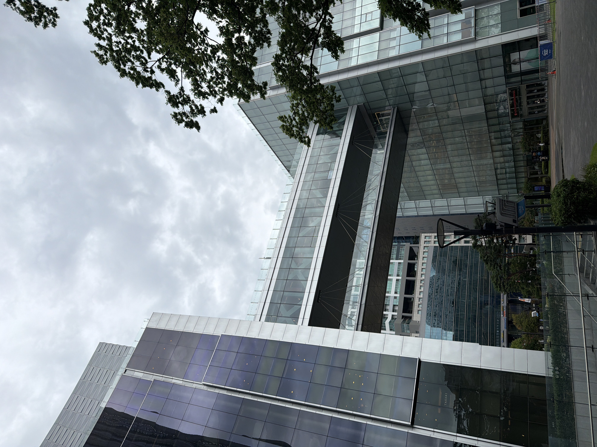 The image depicts a modern architectural scene in the city of Seoul, South Korea. The primary focus is on a large, multi-story building with a distinctive glass facade and an elevated skybridge connecting two sections of the structure. The skybridge appears to be a contemporary design element, enclosed in glass, offering a view of the surrounding urban landscape.
The building's exterior is characterized by sleek, reflective glass panels, which create a sense of transparency and modernity. The architectural style suggests a commercial or corporate complex, possibly housing offices or retail spaces. The skybridge is suspended between two parts of the building, emphasizing its connectivity and dynamic design.
In the foreground, there is a paved area, likely a plaza or walkway, with some landscaping visible. Trees with green foliage are present, adding a touch of nature to the urban setting. The sky is overcast, with a cloudy sky contributing to a diffused lighting condition. A street lamp is visible on the right side of the image, indicating an urban environment.
The overall impression is one of a well-designed, contemporary urban space in Seoul, showcasing modern architecture and urban planning. The image captures a moment of stillness, with no prominent human activity. The reflective surfaces of the building, combined with the cloudy sky, create a somewhat muted yet striking visual.