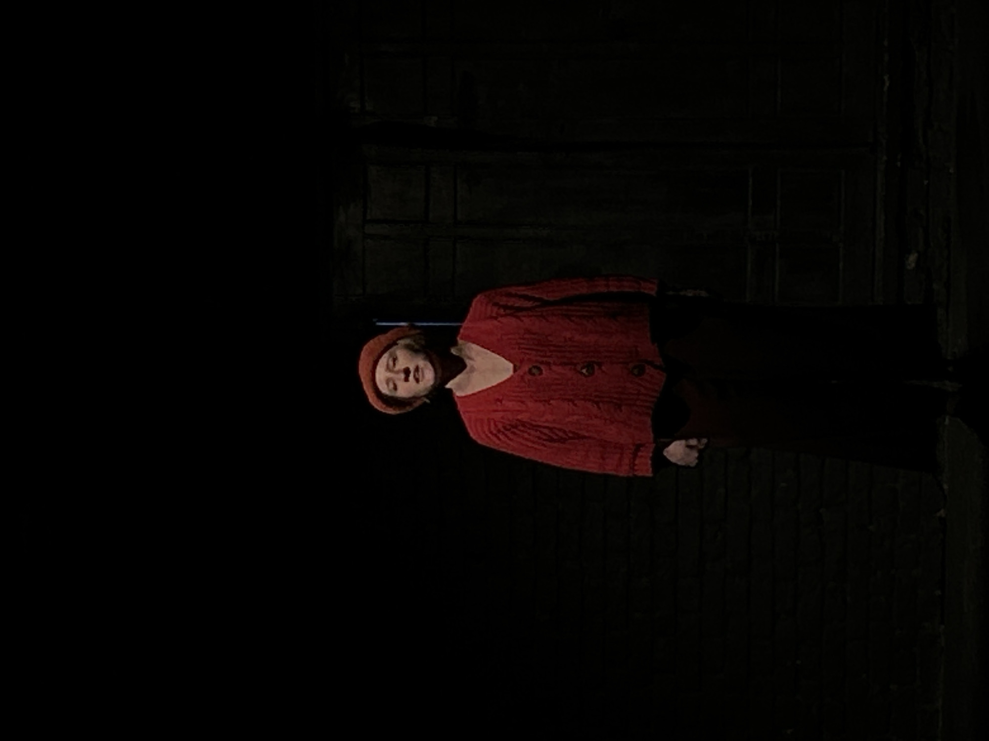 The image depicts a person wearing a red beret and a red cardigan, positioned in front of a dark, boarded-up doorway or wall. The location is a dimly lit area, with the foreground mostly dark and the background providing minimal illumination of the person and the wooden structure behind. The orientation of the image is rotated, with the person and wall appearing sideways. The setting suggests an urban alleyway or similar exterior location.