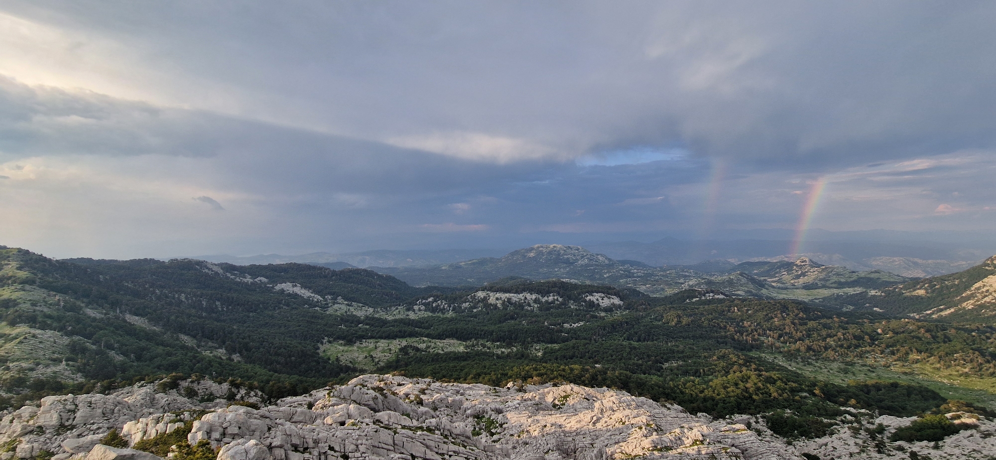 The image is a panoramic landscape view from a rocky, elevated vantage point overlooking a mountainous region. The foreground is dominated by rough, light-colored rock formations, indicative of a karstic landscape. Beyond the foreground, rolling hills covered in dense, dark green forest stretch into the distance. In the mid-ground, there are more rounded mountains, some with visible rocky outcrops.
The sky is dramatic, filled with heavy, grey clouds that appear to be breaking apart in places, allowing shafts of light to pass through. A faint rainbow is visible on the right side of the image, arcing across the sky and disappearing behind a distant mountain range. The overall lighting suggests it is either early morning or late afternoon, with a mix of shadow and illumination across the landscape.
The location is identified as Ubla, Bosnia and Herzegovina, a mountainous area known for its natural beauty. The scene captures a moment of transition, with the moody sky and the presence of a rainbow hinting at recent or impending rain. There are no people or man-made structures immediately visible in the frame, emphasizing the natural and wild character of the environment. The image conveys a sense of vastness and rugged terrain.