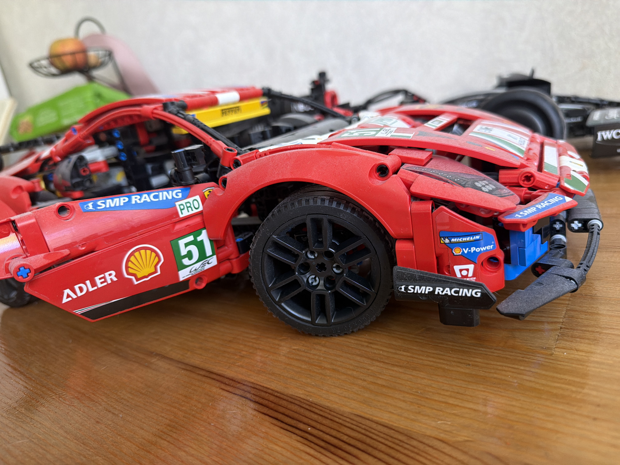 This image shows a red Lego Technic Ferrari race car model resting on a wooden surface. The car is detailed with various stickers representing sponsors and racing numbers, including "SMP Racing", "Adler", "Shell", and the number "51". The car also features a Michelin "V-Power" sticker. The focus is on the front portion of the car, including the wheel, fender, and front spoiler. In the blurred background, another Lego model of a black race car can be partially seen. An apple sits in a wire basket in the background. The location cannot be determined from this image.