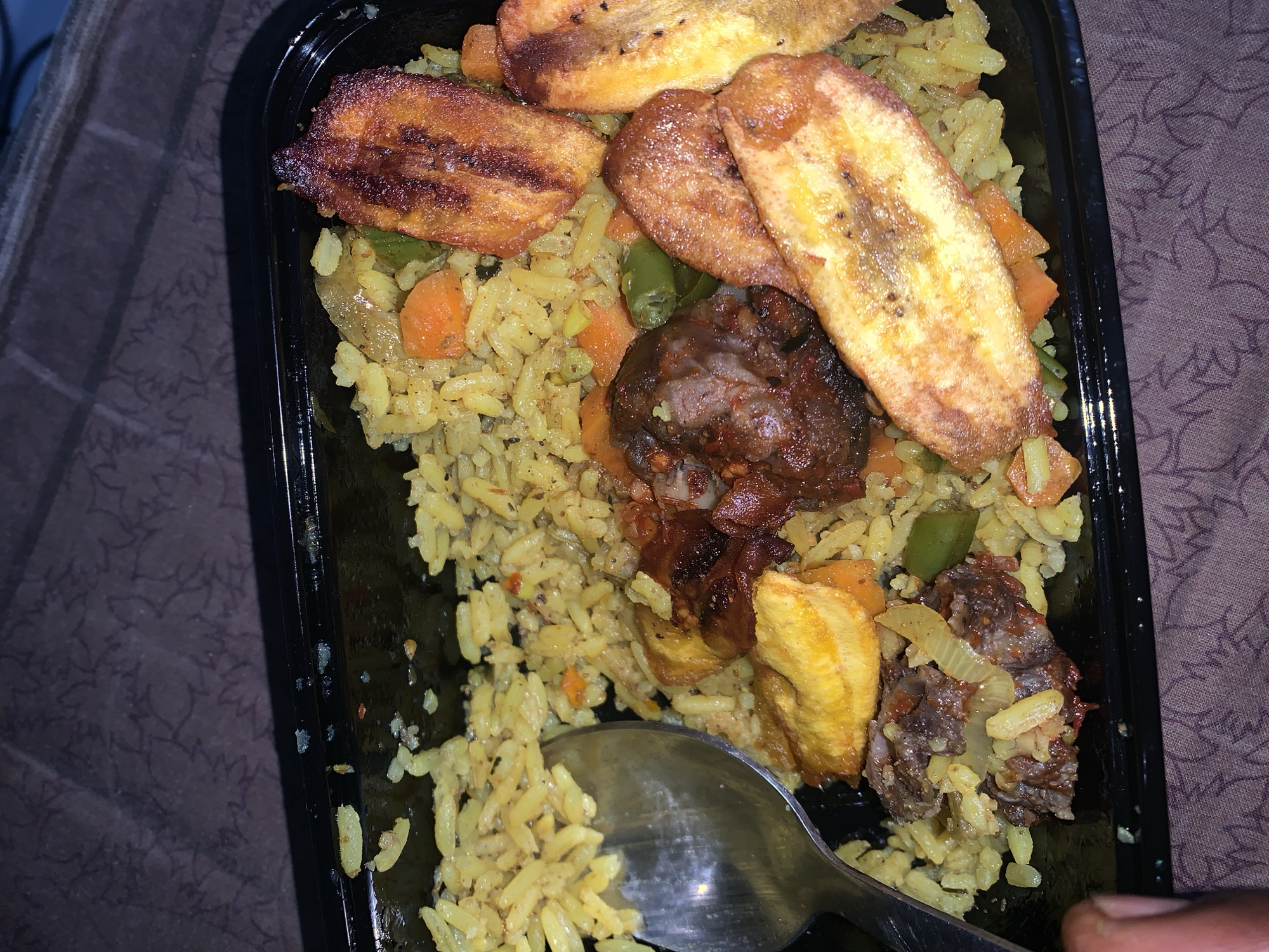 The image shows a black plastic food container filled with yellow rice, fried plantains, and meat. Visible ingredients mixed with the rice include green beans, carrots, and onions. A metal spoon is in the container, partially covered by the rice. The container is resting on a textured surface.
The image doesn't contain the necessary information to locate this image in the city of Rafin Par, Nigeria.
