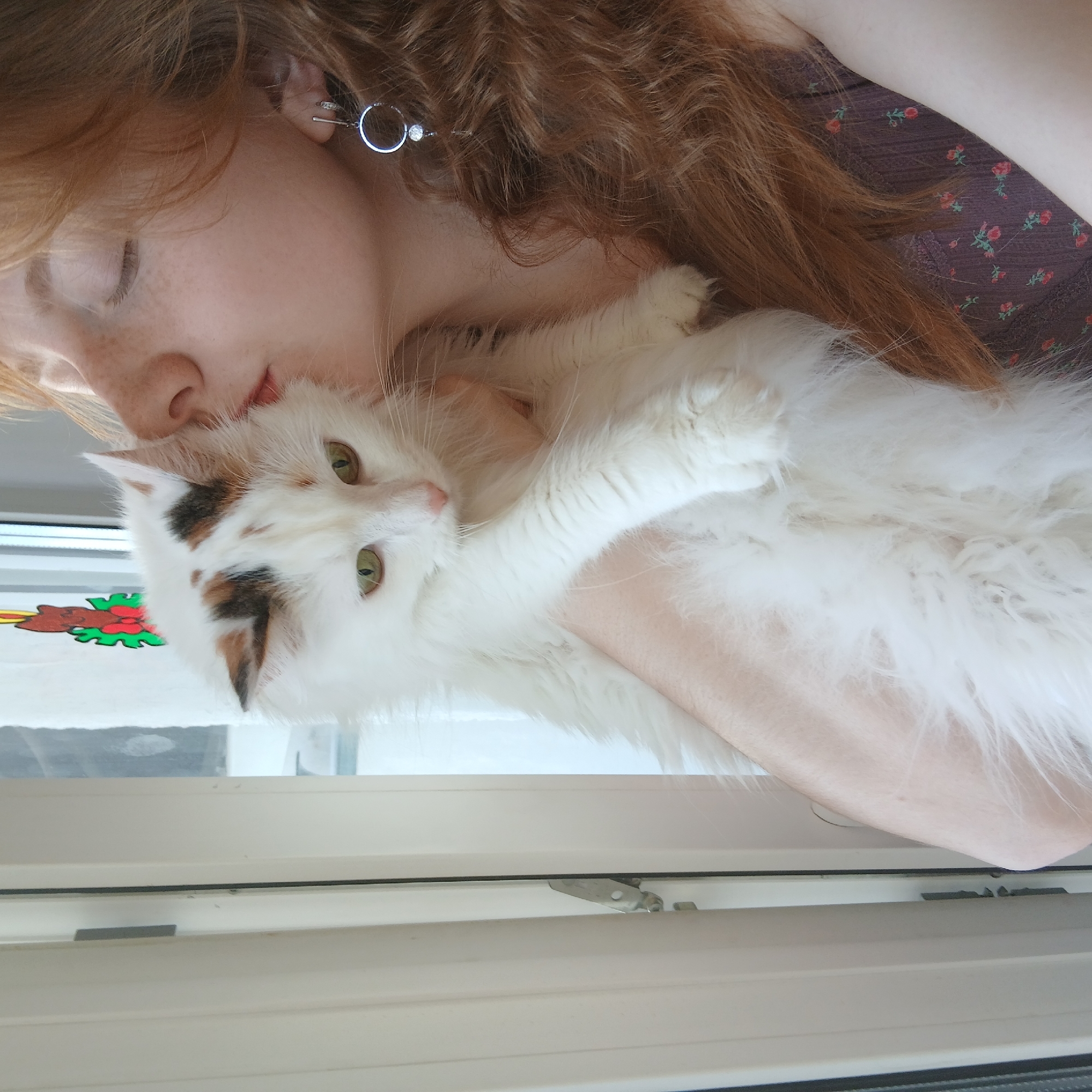 A fair-skinned, red-haired woman with freckles is holding a white, long-haired cat with patches of brown and black fur. The woman's eyes are closed as she nuzzles the cat's head. She is wearing a patterned top, and a silver hoop earring is visible in her left ear. The cat's green eyes are open. They appear to be near a window, possibly indoors. The image orientation is sideways. This location cannot be verified to be in the city of Yekaterinburg, Russia.