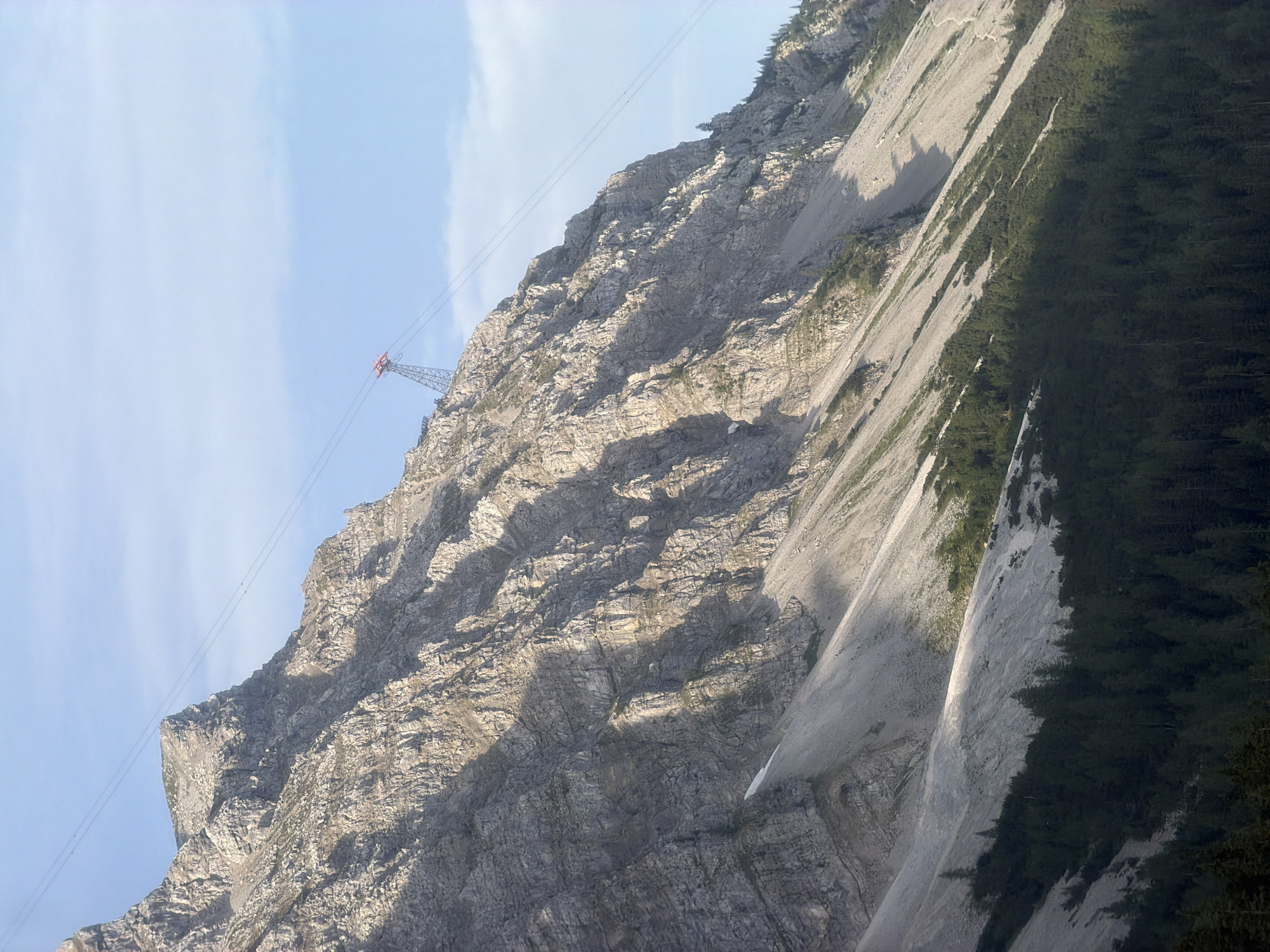 This image depicts a mountainous landscape near Grainau, Germany, featuring the Zugspitze cable car. The scene is dominated by a steep, rocky mountain face, characterized by rugged textures, striations, and shadows cast by the sunlight. The lower slopes are covered with dark green coniferous trees, creating a distinct contrast with the lighter colored mountain scree and rock formations above.
A prominent feature is the red and silver pylon of the Zugspitze cable car, situated on the upper part of the mountain slope. Two parallel steel cables extend from the pylon diagonally upwards and across the frame, disappearing towards the left edge of the image. The sky is a clear, light blue with faint wisps of clouds. The overall impression is one of a vast, natural landscape with a man-made structure integrated into it, likely a scenic viewpoint or transportation hub.