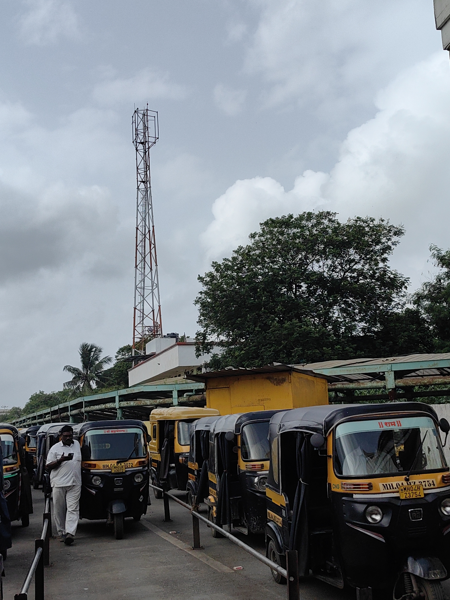 The image depicts a row of auto-rickshaws parked along a road, likely a bus or auto stand, in Mira-Bhayander, India. A man in a white shirt and pants is walking next to one of the auto-rickshaws, looking at his phone. The sky is overcast. A tall telecommunications tower is visible in the background, along with some trees and a yellow building. The auto-rickshaws are mostly black and yellow, with some visible license plates. The scene appears to be during the daytime.