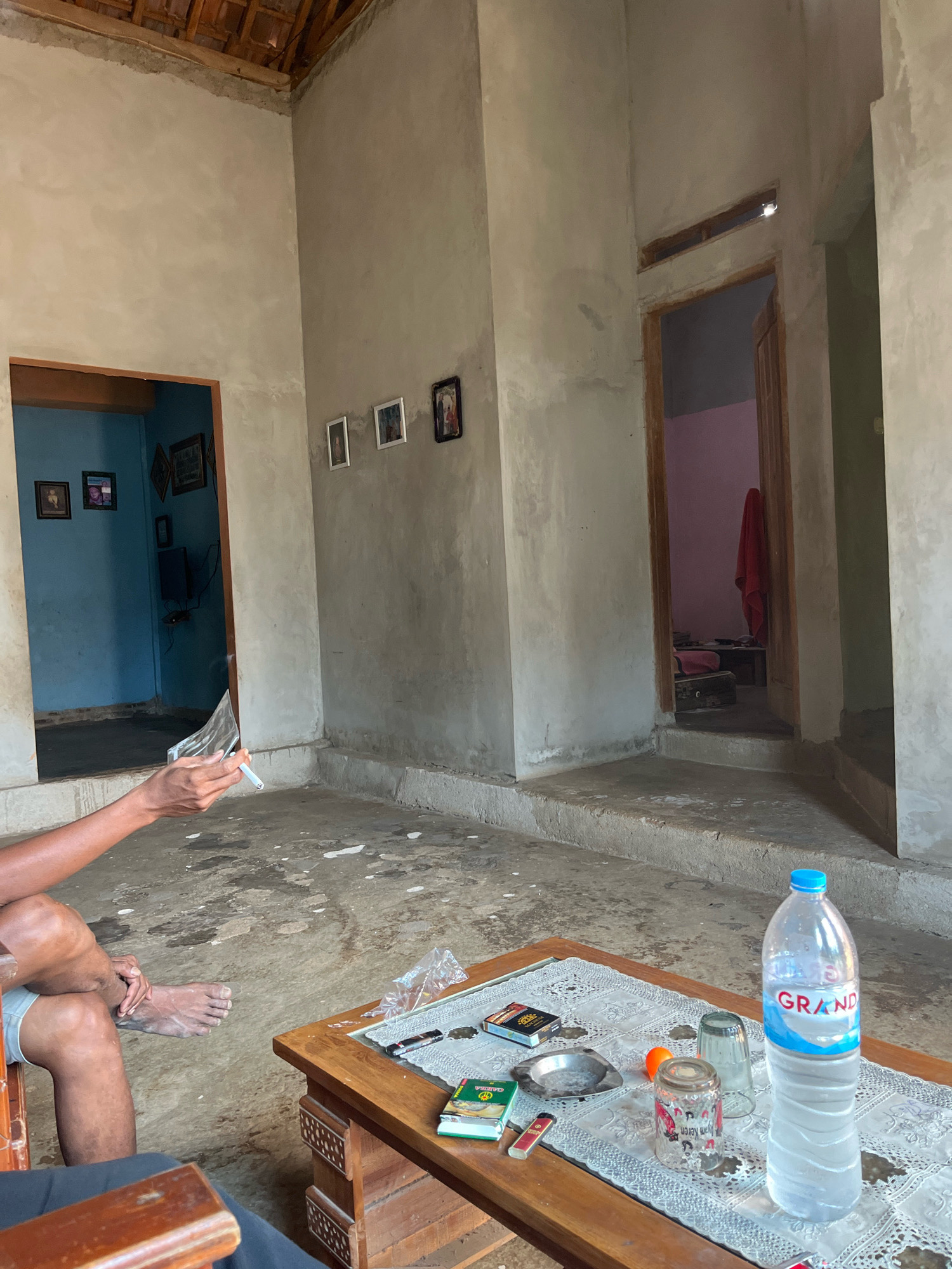 The image depicts an interior scene of a modestly furnished dwelling, likely a home, in Sinar Mulyo, Indonesia. The walls are unpainted concrete, and the ceiling appears to be constructed of wooden beams and tiles. The flooring is a rough, stained concrete. Two doorways are visible, one to the left leading into a room painted blue and containing a television and framed pictures, and another to the right opening into a room with pink walls and a red cloth hanging.
In the foreground, a wooden coffee table is set with items including a bottle of "GRAND" brand water, a glass, a small ashtray, two packs of cigarettes, a lighter, and a small book. A person, partially visible sitting in a chair on the left, is holding a cigarette in their right hand and appears to be interacting with a piece of transparent material. Their legs are crossed, and their bare feet are visible on the floor. Another person is seated in a chair in the lower foreground, partially obscured. The overall impression is a casual, lived-in space.