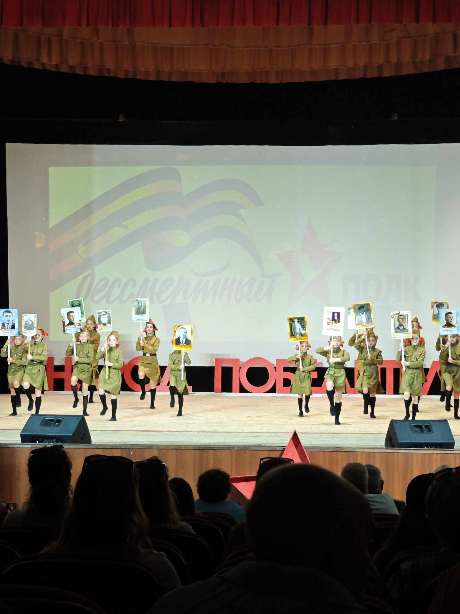 Captured in Марковский, Russia, the image depicts a stage performance where a group of young girls, dressed in olive green uniforms and black knee-high socks, are engaged in a choreographed dance. Each girl carries a rectangular sign displaying a black and white portrait of a person, potentially commemorating historical figures. Behind the dancers, a large screen projects a digital image containing Russian text and a design resembling ribbons and a star, possibly related to a historical event. The stage is decorated with oversized red Cyrillic letters, spelling out "Победа" (Victory). In the foreground, the backs of the audience members' heads are visible, suggesting a live performance setting. Two speakers are visible on the front of the stage. A red star prop is also visible on the stage, partially obscured by a person in the audience. The curtains above the stage are dark red.