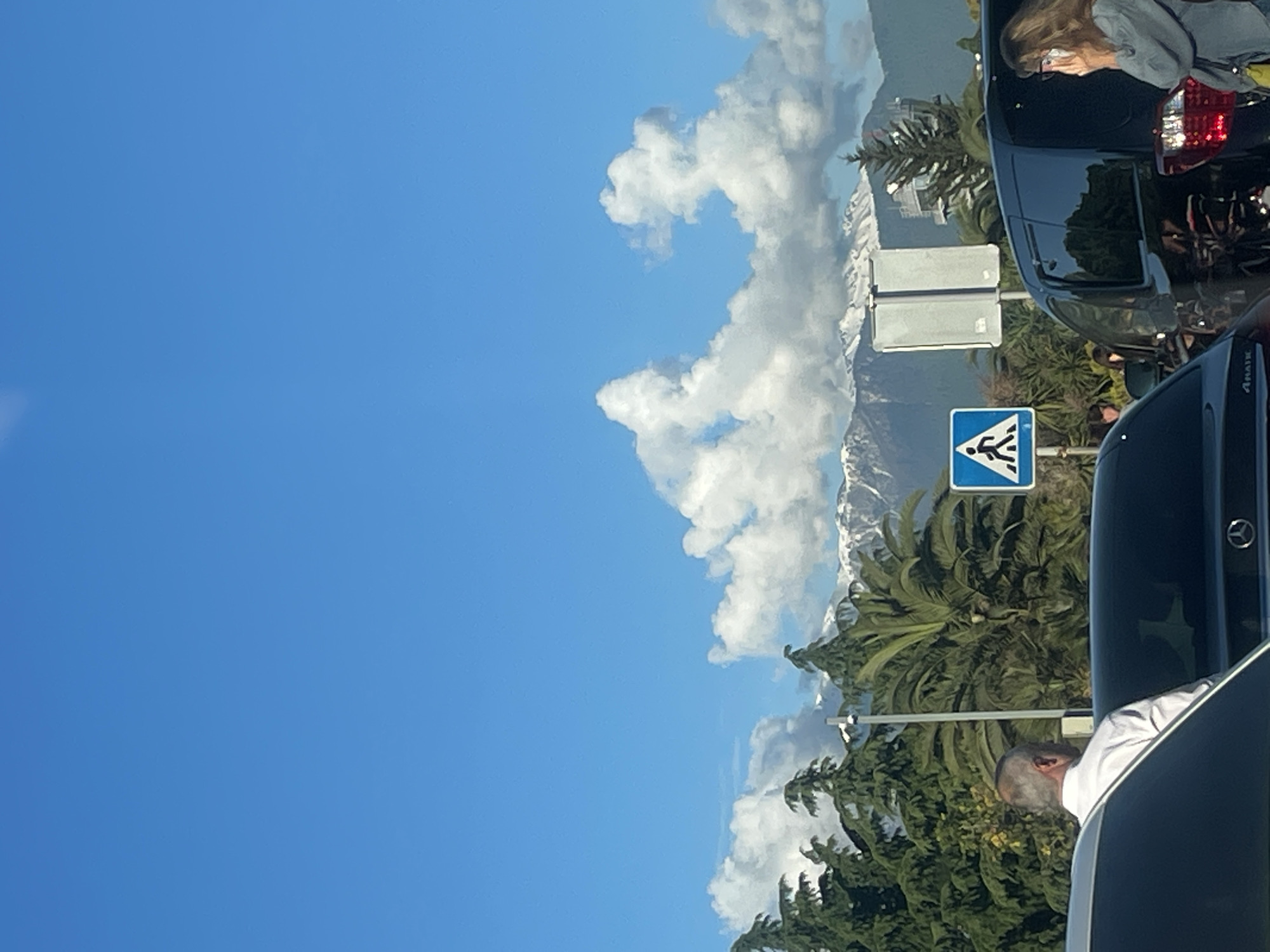 The image shows a daytime urban scene, likely taken from a vehicle. A clear blue sky dominates the left side of the image. Fluffy white clouds partially obscure snow-capped mountains in the background. Closer to the foreground, green palm trees and other foliage are visible. Street signs, including a pedestrian crossing sign, are present. Several vehicles are visible in a traffic jam, including portions of the car from which the image was taken. The partial view of a woman with a mask inside one vehicle and a man's head visible in another. Based on the palm trees and mountains and that there are cars in the foreground, this may be the city of Sochi, Russia.
