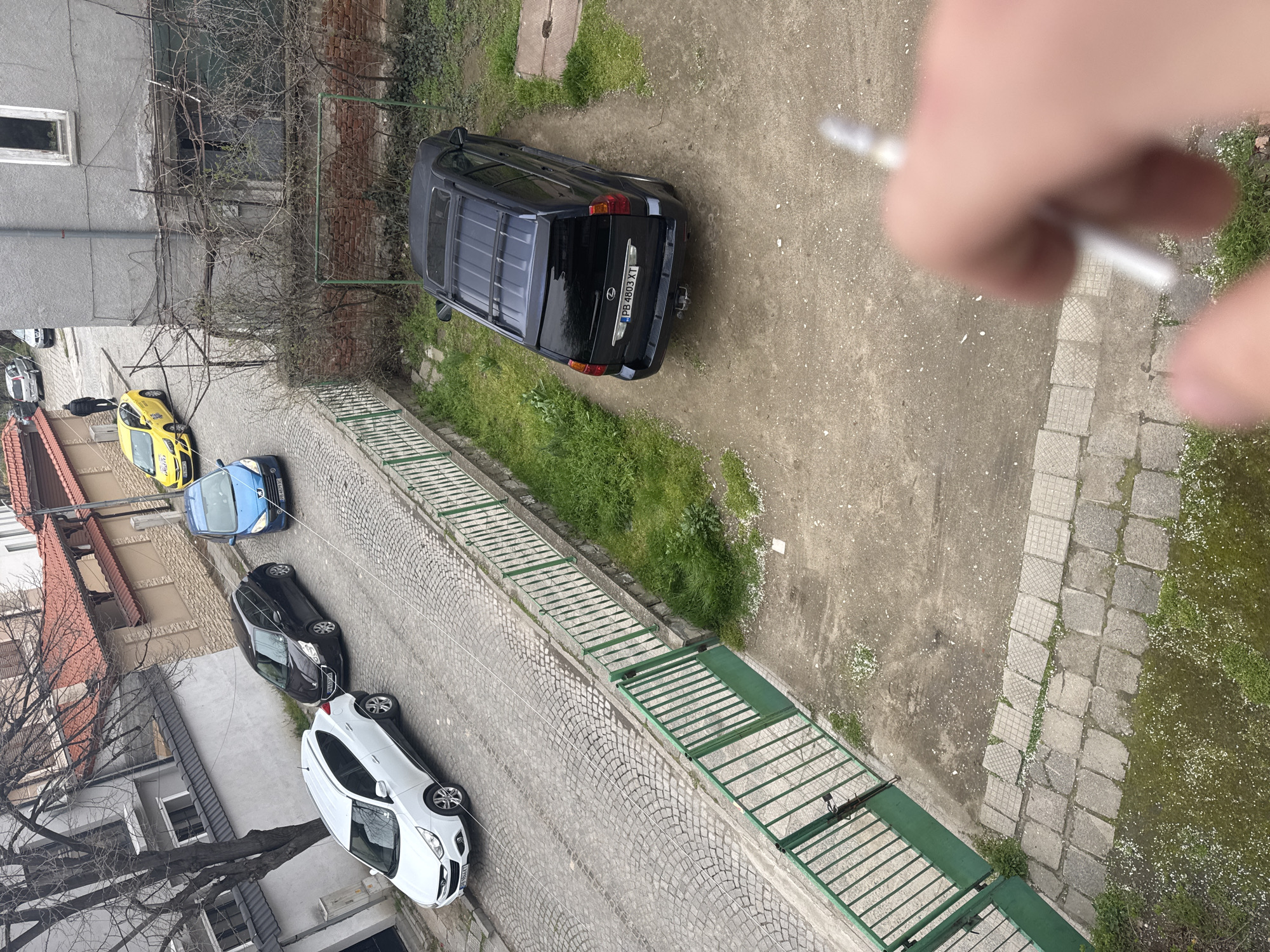 A high-angle photograph captures an outdoor scene in Plovdiv, Bulgaria. The image is dominated by several parked cars: a dark blue SUV, a bright yellow taxi, a blue compact car, a dark grey sedan, and a white hatchback. They are parked along a cobblestone street, adjacent to a green metal fence that separates the street from a grassy area. The architecture includes buildings with pitched roofs and tiled rooftops, typical of the region. A partially visible hand holding an object obscures the upper right portion of the image, adding an element of perspective from the photographer's viewpoint.