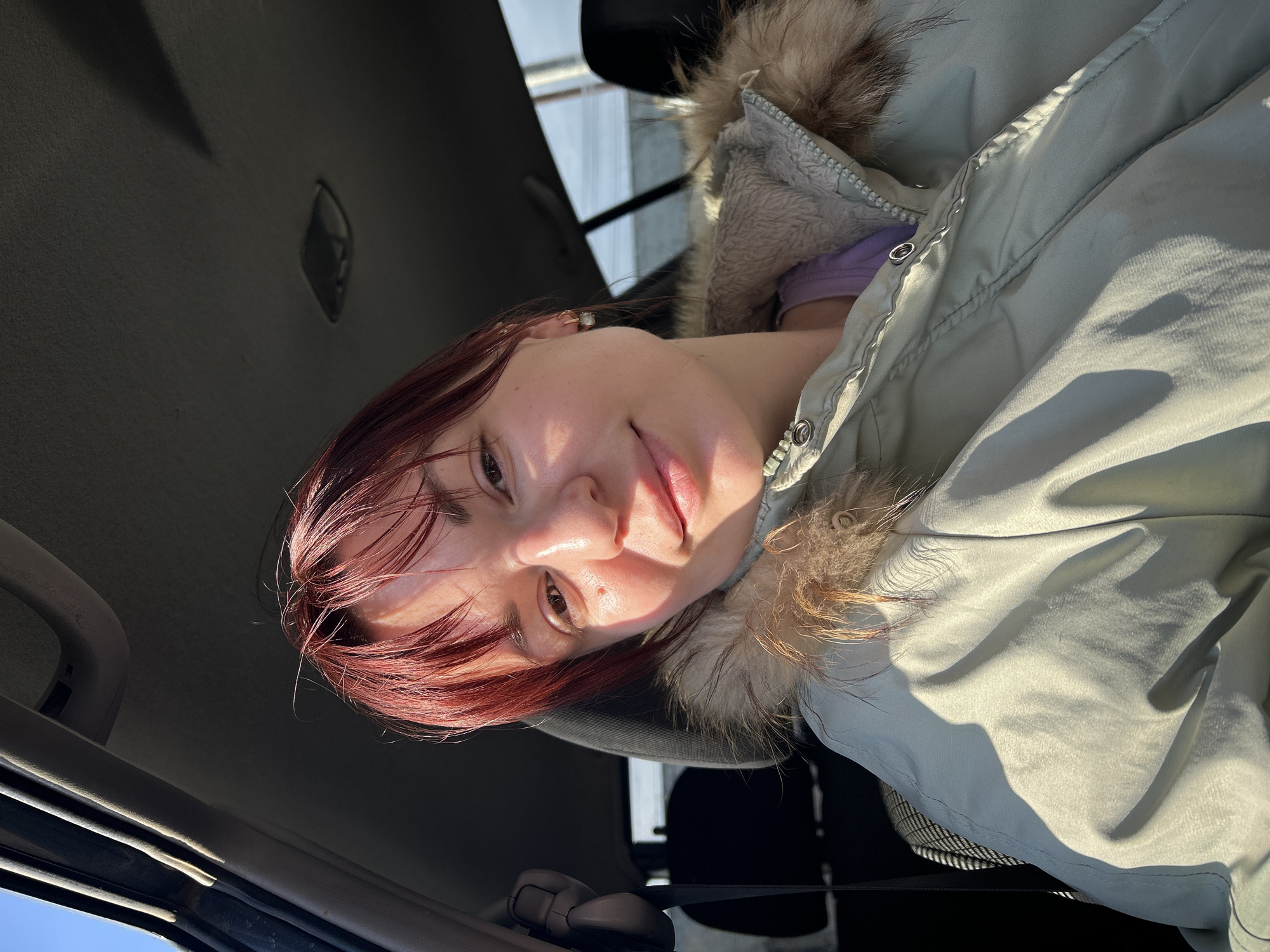 A woman with auburn hair, wearing a light green jacket with a fur-lined hood and a purple shirt underneath, is sitting in a car. The interior of the car is dark, with the roof and sun visor visible. Outside, the sky is bright. The location cannot be determined to be Chaikovsky, Russia from this image.