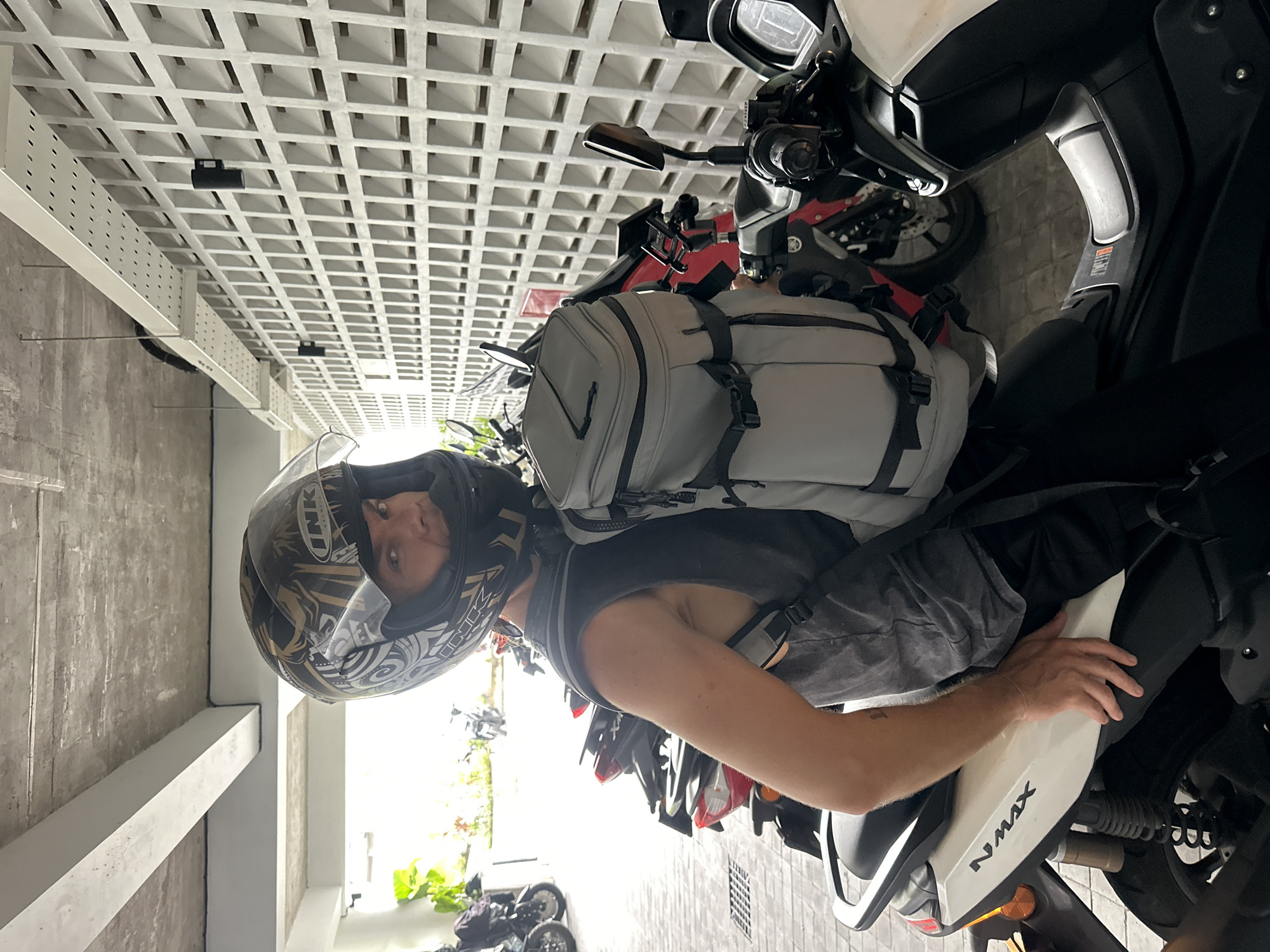 A person wearing a dark helmet and a gray backpack is sitting on a white NMAX motorcycle in an indoor parking area. The person appears to be looking directly at the camera. Behind the motorcycle and person, other motorcycles are parked in a row. The ceiling is composed of a grid-like structure of white squares. The location is Mengwi, Indonesia.