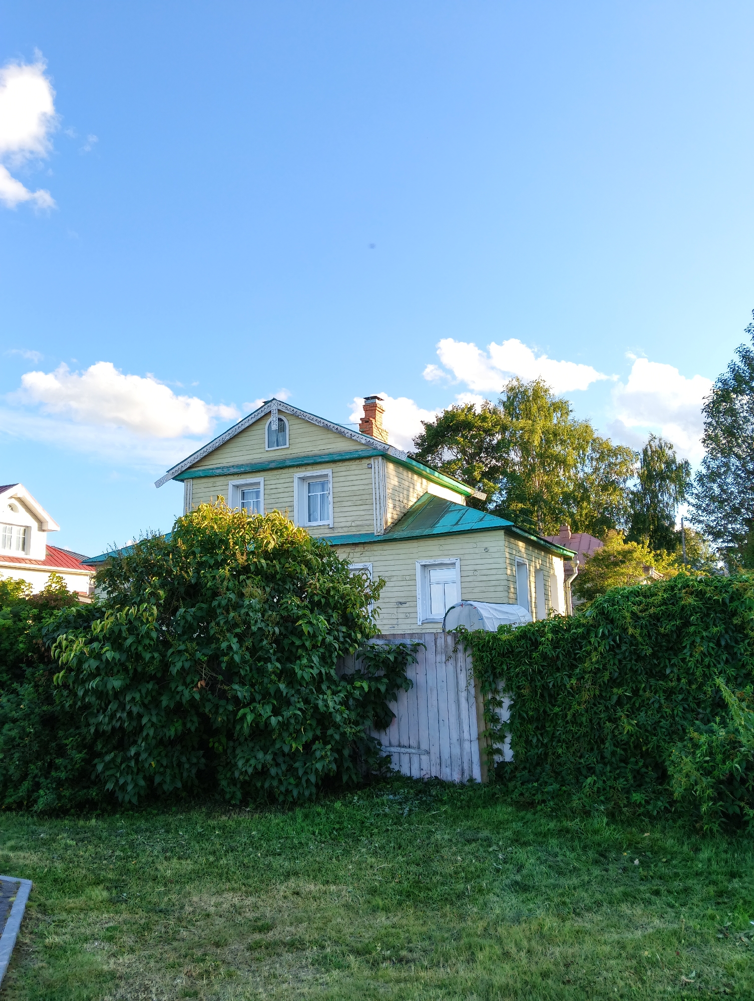 A pale yellow, two-story wooden house with a teal roof and a red brick chimney stands behind a white wooden fence. The house has white-trimmed windows, and decorative white trim under the eaves. The fence is partially covered in green ivy. In the foreground, a patch of green grass is visible, with a dark gray concrete path on the left. The sky above is blue with scattered white clouds. The image appears to be taken in a residential area of Petrozavodsk, Russia, likely during the daytime.