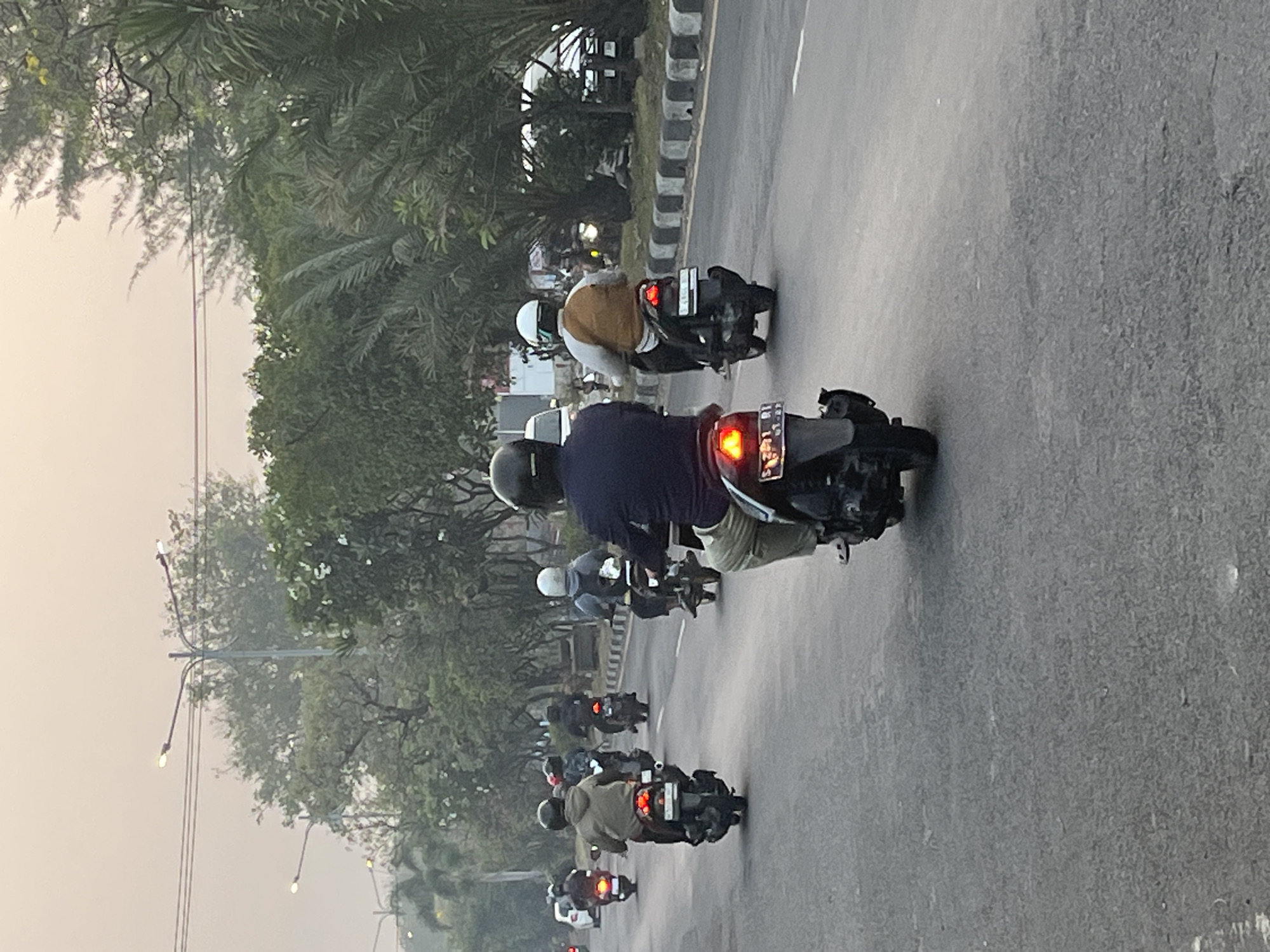 The image shows a group of people riding motorcycles on a street in Ngaban, Indonesia. The weather appears overcast or hazy, and the sun is low in the sky, suggesting it is either early morning or late afternoon. The motorcycles are moving in the same direction on a paved road. The riders are wearing helmets and various casual clothing. Trees and palm trees line the side of the road, and streetlights are visible in the distance. The overall impression is of a typical urban or suburban street scene in Indonesia during a time of reduced daylight.