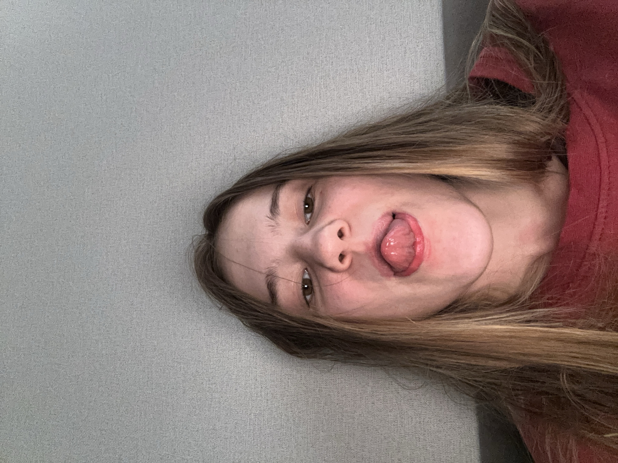 The image is a close-up, selfie-style photograph of a young person with long, blonde-brown hair, sticking their tongue out. The person is wearing a red shirt. The background is a plain, textured gray wall. The lighting appears to be artificial and is coming from the front. The location is not identifiable from the image itself, and therefore any information about Vsevolozhsk, Russia is speculative.
