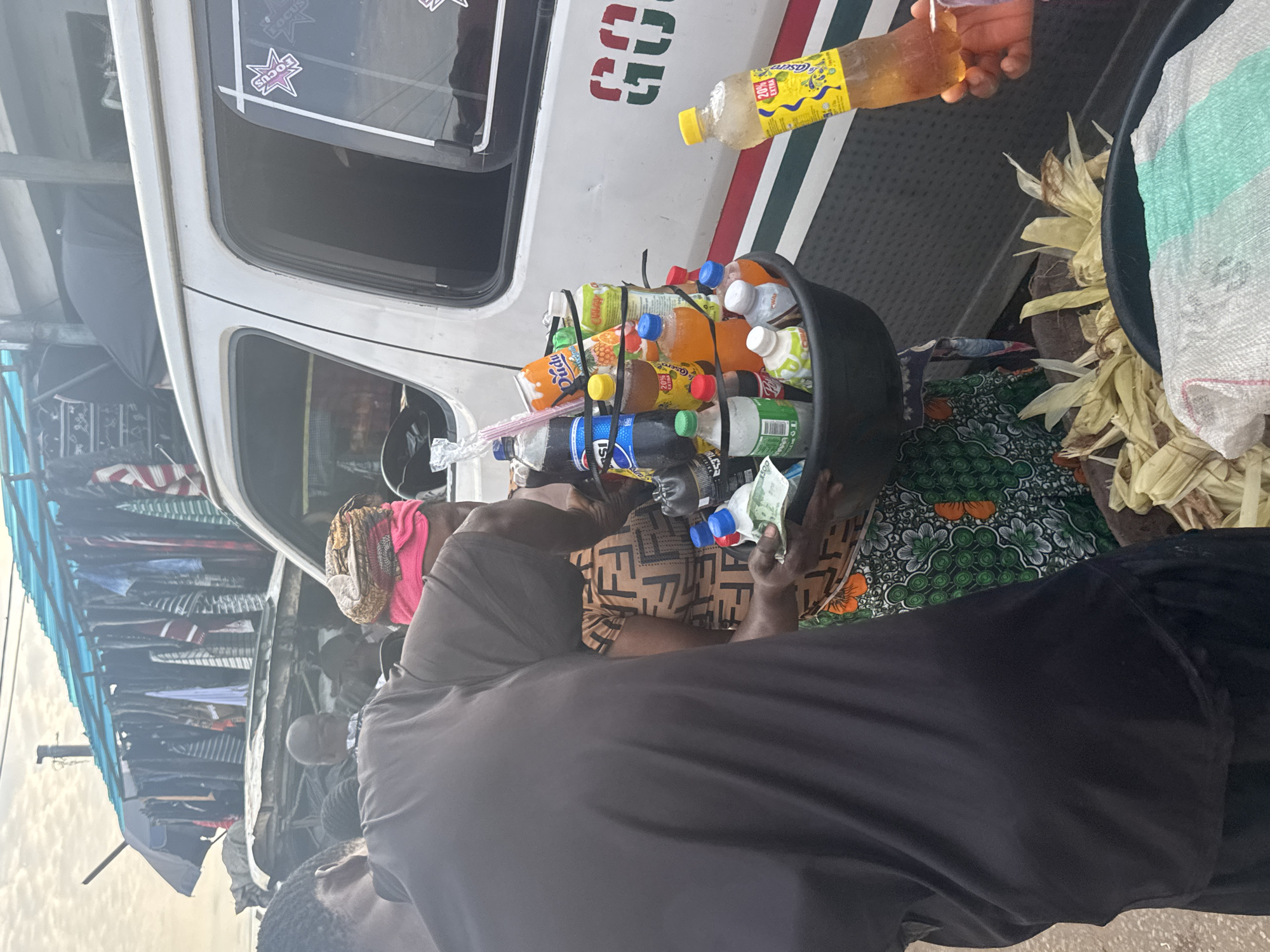 In the city of Onitsha, Nigeria, a street vendor is offering a selection of bottled drinks from a dark basin to a customer. The vendor, a woman wearing a colorful patterned dress and a pink headscarf, stands beside a white van. She holds a few banknotes in her hand, suggesting a transaction is underway. Beside her, a person in a dark t-shirt appears to be the customer, their back turned to the camera. The van has green and red stripes along its side, and the word "GO" is partially visible in red lettering. In the background, a makeshift stall displays an assortment of clothing items hanging from racks under a blue tarp. The scene appears to be an outdoor market, characteristic of busy urban areas in Nigeria.