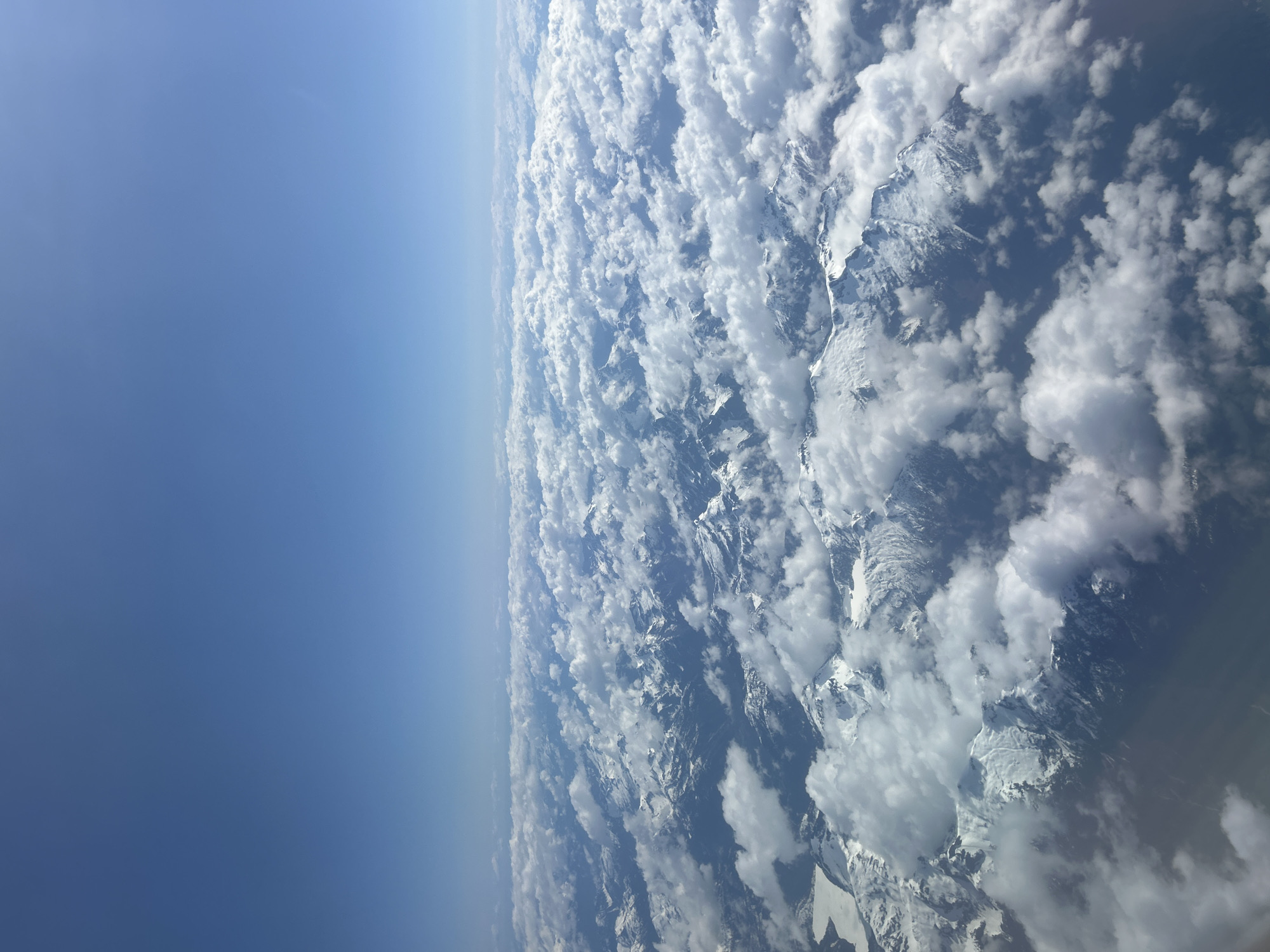 This media file presents a high-altitude aerial view, likely captured from an aircraft, showcasing a vast mountainous landscape partially covered by clouds under a clear blue sky. The image is oriented vertically.
On the left side, a broad expanse of brilliant blue sky dominates, transitioning smoothly from a deep azure at the top to a lighter shade closer to the distant, hazy horizon line, which runs diagonally across the middle of the frame. This clear sky suggests a high altitude.
The right side of the image features rugged, snow-capped mountain ranges intermingled with dense formations of white cumulus clouds. Numerous peaks, dusted with snow and ice, are visible through breaks in the clouds, their textured surfaces showing a mix of bright sunlight and deep shadow. The clouds vary in density and form, appearing both as widespread layers and individual puffs, often nestled among the mountain contours or towering above them. This creates a striking contrast between the bright white clouds and the darker, shadowed mountain faces.
The scene depicts a bright daytime, likely mid-day, with strong, direct illumination. Weather conditions indicate clear skies at very high altitudes, while the mountain regions are partly cloudy. Given the provided context of Kelowna, Canada, the view likely showcases a portion of the extensive mountain ranges in British Columbia, such as the Canadian Rockies or the Coast Mountains. There is no identifiable text visible within the image.