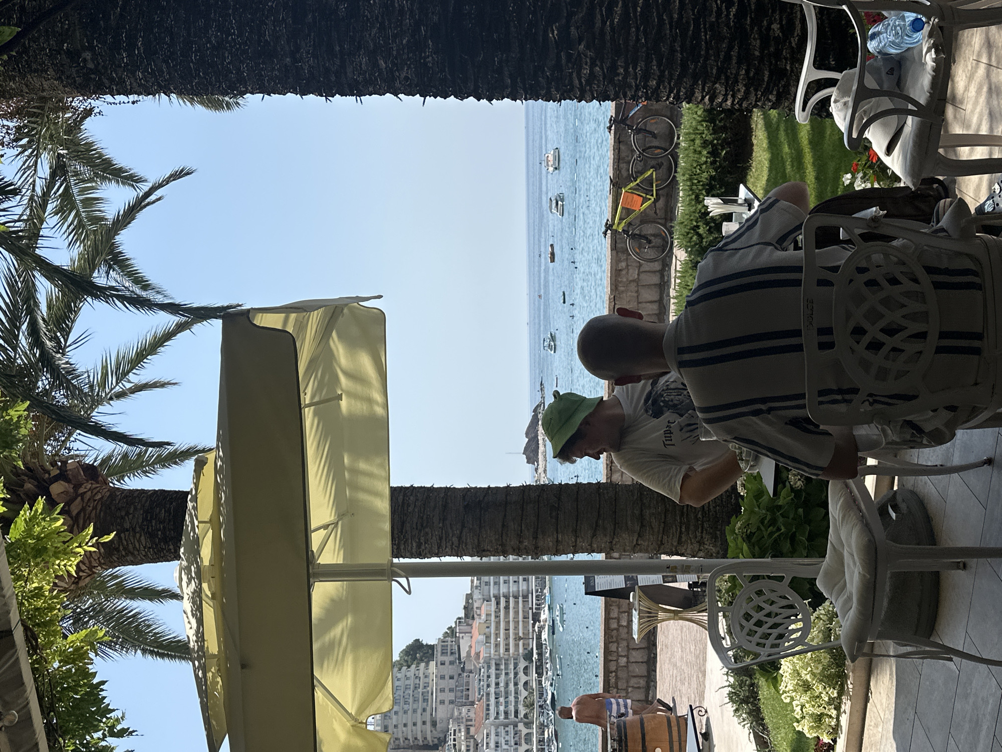 The image captures a bright, sunny outdoor scene at a waterfront cafe in Rafailovići, Montenegro. In the foreground, two men are seated at tables, facing away from the camera. One man wears a striped shirt, and the other wears a white t-shirt and a green hat. White metal chairs with cushions are placed around the tables. To the left, a man in shorts walks away from the camera.
The cafe area overlooks a blue sea dotted with several small boats. In the background, a hillside is covered with buildings, suggesting the urban setting of Rafailovići. Tall palm trees, one with a thick, textured trunk, frame the view, with their fronds casting shadows on the scene. Bicycles are parked on a paved area behind the cafe seating. The overall atmosphere is relaxed and vacation-like, typical of a coastal town.