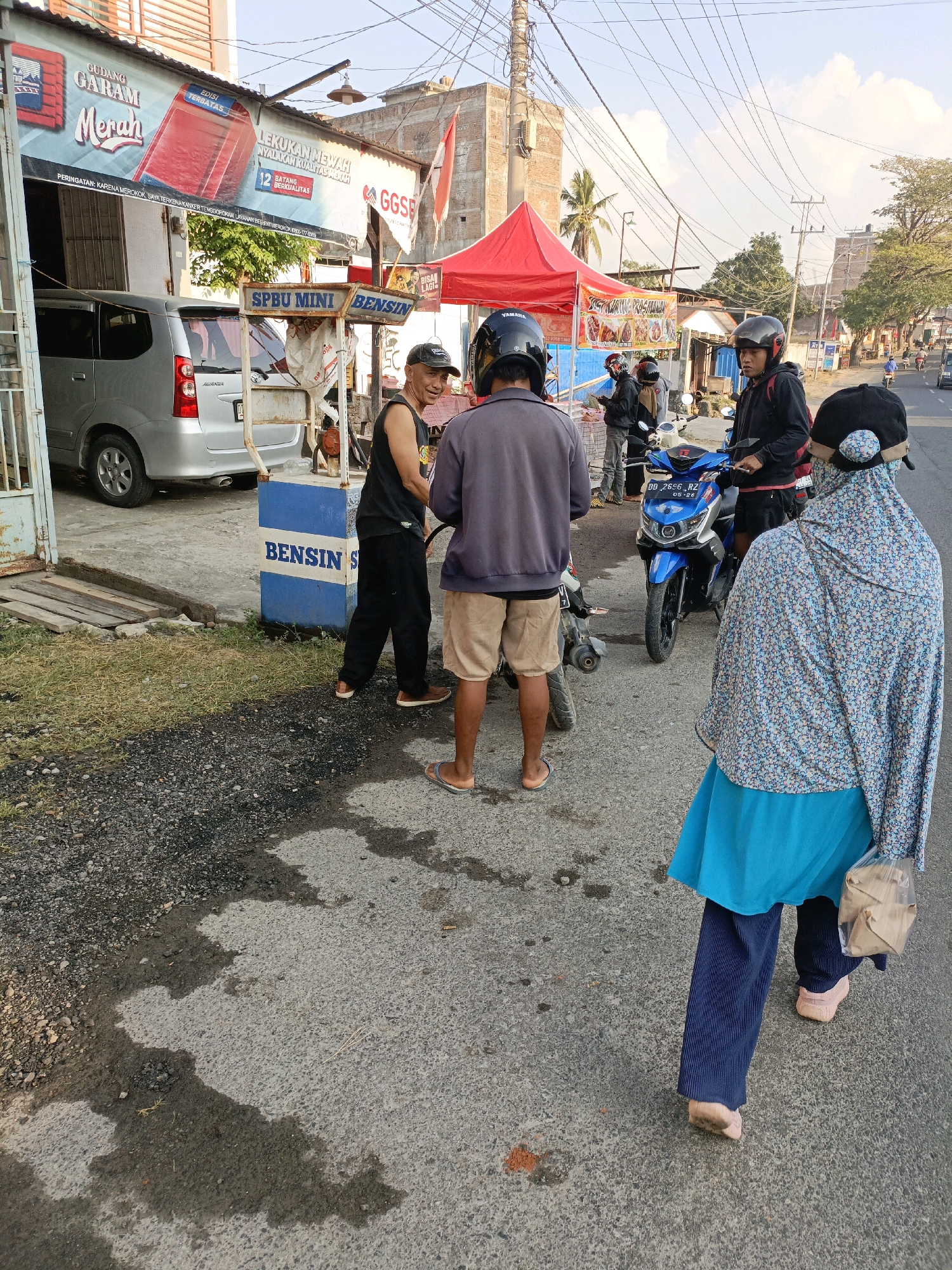 The image captures a street scene in Bulukumba, Indonesia, likely near a small roadside shop. A silver minivan is parked on the left side of the frame. In front of a building with a sign that reads "GUDANG GARAM MERAH" and other Indonesian text, a blue and white gasoline pump labeled "SPBU MINI BENSIN" is visible.
Several individuals are present around the gas pump and on the street. Two men, one wearing a black tank top and a cap, and another in a grey jacket and beige shorts, are interacting near the pump. A third man in a black jacket and helmet is standing by a blue motorcycle. Further down the street, a person wearing a blue hijab and a floral pattern shirt, and blue trousers, walks away from the camera, carrying a light brown bag. Another motorcycle is parked next to the blue one. A red tent is set up behind the gas pump, possibly for a vendor. The environment appears to be a typical roadside setting in Indonesia with a paved road, gravel on the side, and overhead electrical wires. The sky is partly cloudy, suggesting daytime.