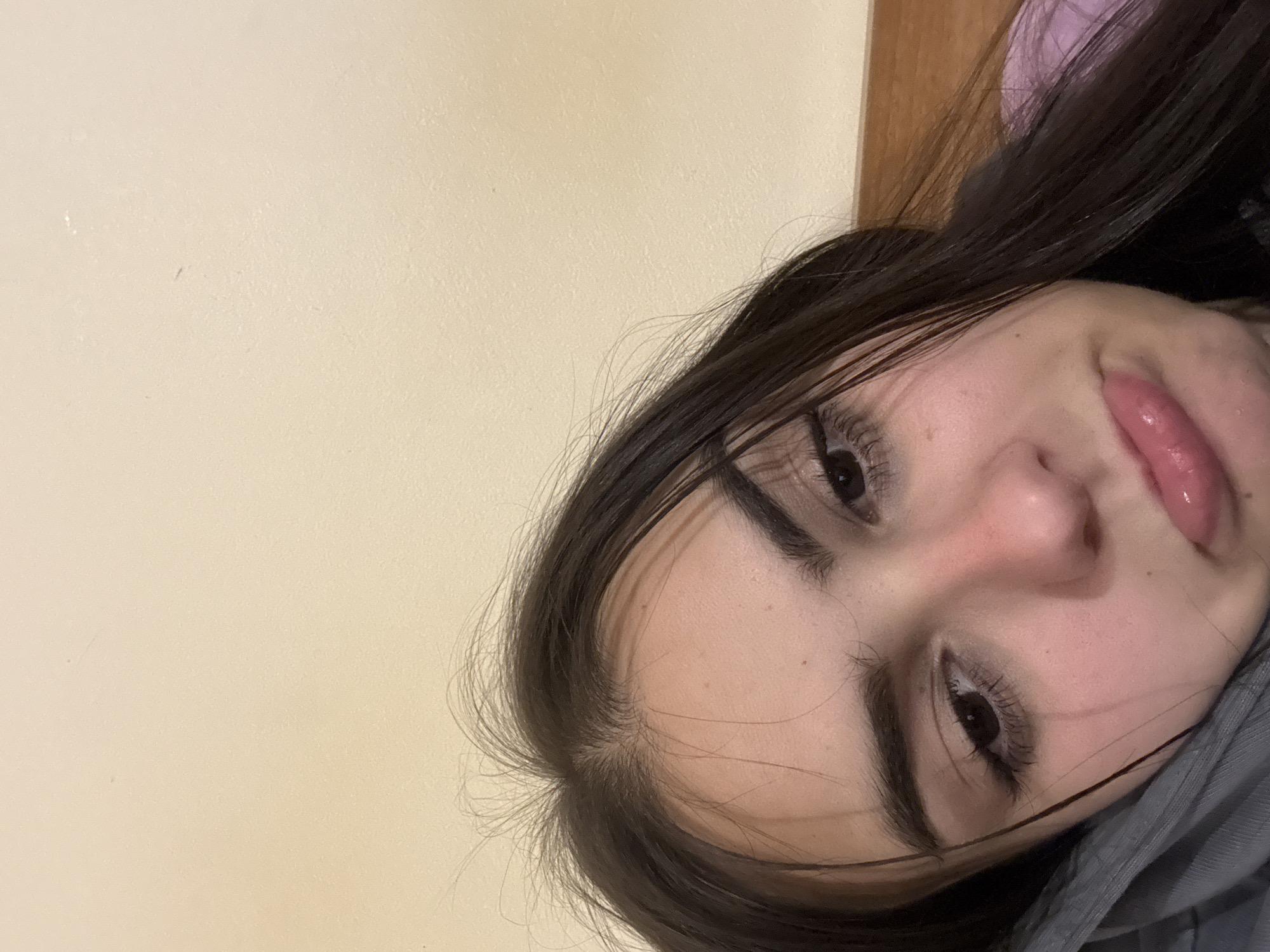 A close-up, slightly angled portrait of a young woman with dark hair. Her face is the primary subject, featuring her eyes, nose, and partially open mouth with glossy lips. The background is a plain, light-colored wall. Part of a gray garment is visible in the lower right corner, suggesting she might be wearing a jacket or sweatshirt. There is no information to suggest this is in Blagoevgrad, Bulgaria.