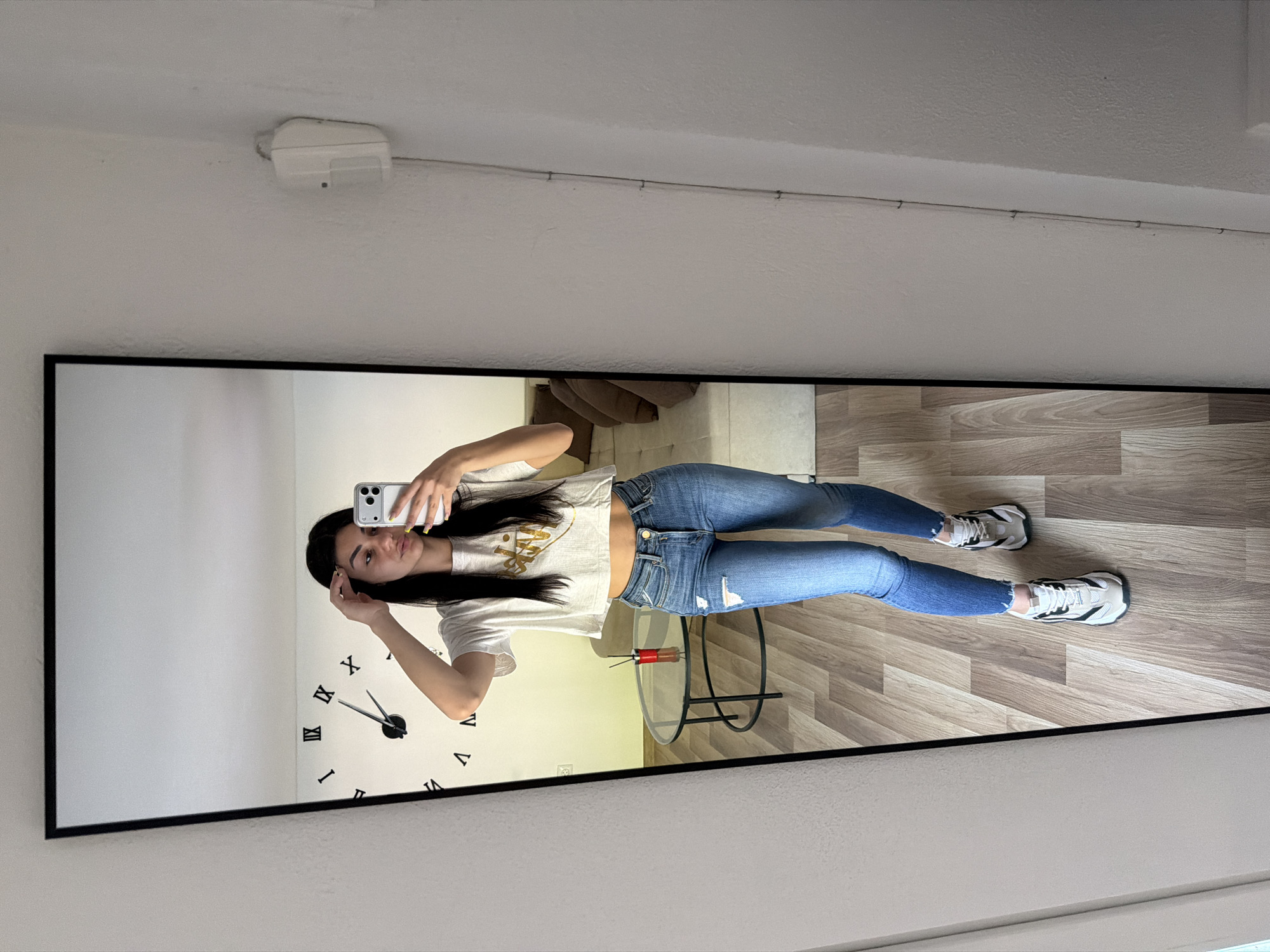 A full-length mirror hanging on a white wall shows a reflection of a young woman taking a selfie. The woman is wearing a white t-shirt, blue jeans, and sneakers. Her dark hair is long and partially covers her face. She holds a cell phone in her right hand, seemingly capturing her reflection. Behind her, a wall clock is visible on the left. A glass-topped table with black legs stands near her waist. The floor has light brown wood-like panels arranged in a diagonal pattern. In the background, partly visible through the reflection, is a tan colored sofa. A white smoke detector is mounted on the ceiling above the mirror. A wire extends from the detector and along the wall, secured by small fasteners.