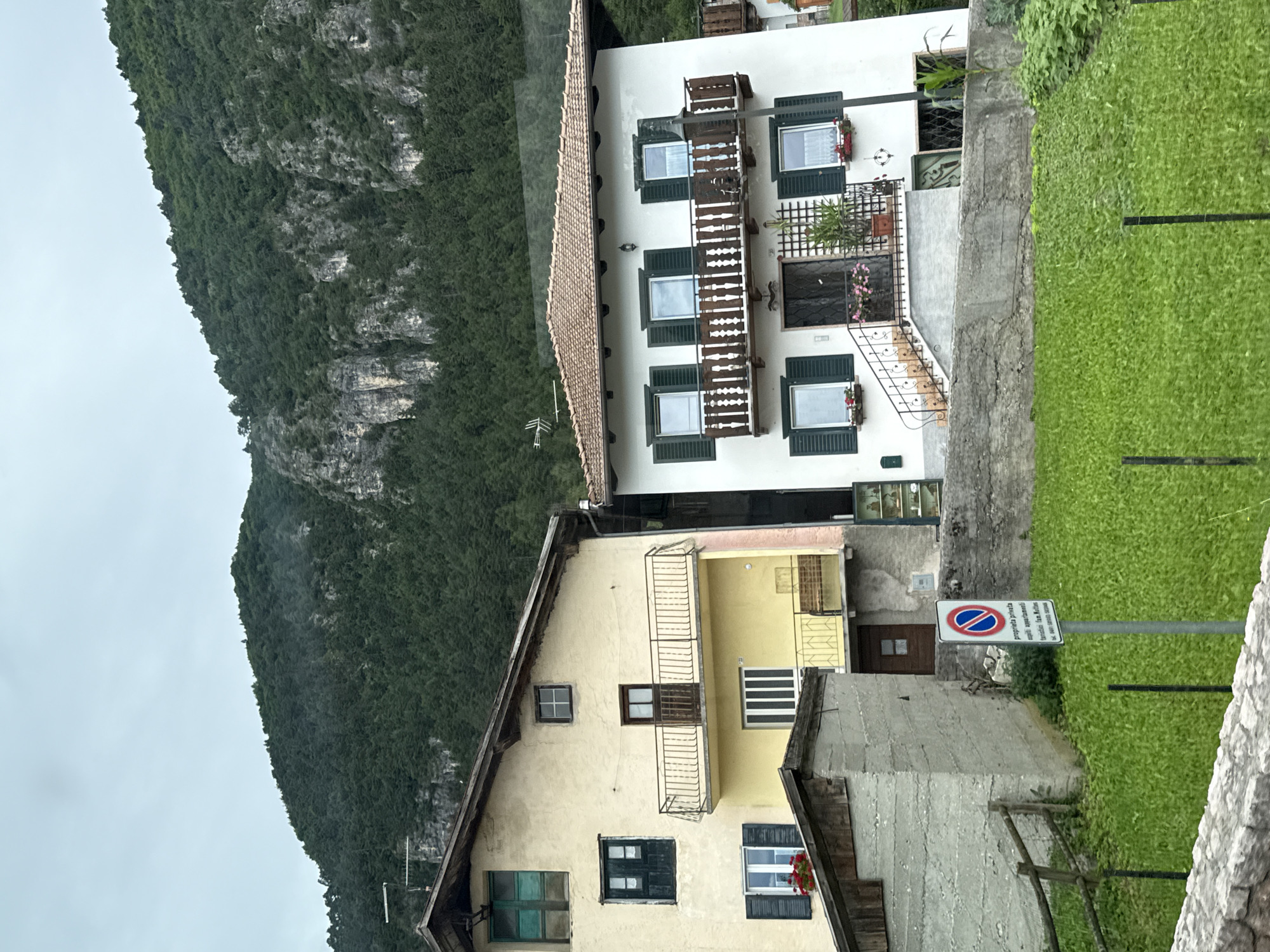 The image depicts two houses situated on a hillside in Fai della Paganella, Italy, with a densely forested mountain slope dominating the background under an overcast sky. The house on the left is painted a pale yellow and features multiple windows with dark green shutters, some of which are open, revealing the interior. A balcony with a wrought-iron railing is visible on the second story, above a lower yellow-painted section. To the left of this house, a smaller, grey concrete structure with a wooden roof extends towards the foreground.
The house on the right is white with dark green shutters on its windows, similar to the neighboring building. It has a prominent wooden balcony with ornate white railings on the upper floor. A set of stairs with a decorative metal railing leads to the entrance on the ground floor, which has a black-grated door. The roof of this house is covered with terracotta tiles.
In the foreground, a lush green lawn stretches out, bordered by a low stone wall and a wooden fence. A signpost with a circular red and blue symbol and Italian text, indicating "private property" and "authorized parking," stands on the lawn. The overall scene suggests a residential area nestled within a mountainous landscape.