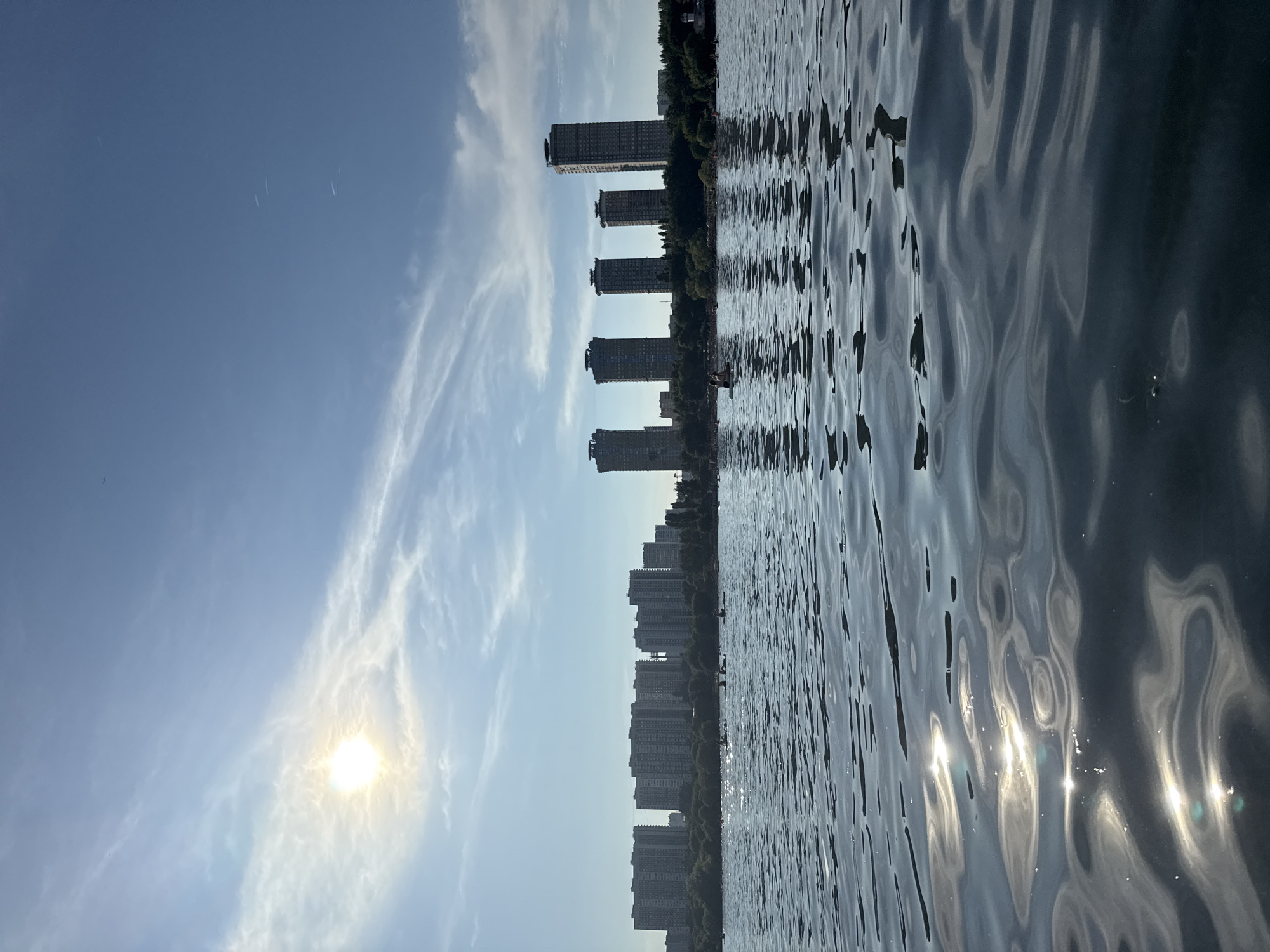 This is a landscape photograph taken from a body of water, possibly a lake or a river, looking towards a cityscape under a bright, partly cloudy sky. The sun is visible in the upper left quadrant, casting a warm glow and creating reflections on the rippling water's surface.
In the mid-ground, a line of buildings stretches across the horizon, ranging from mid-rise apartment complexes to taller, modern skyscrapers. The architecture suggests an urban environment. Trees and greenery are visible between and in front of the buildings, forming a natural buffer.
On the water, there are several small boats, some with people in them, and a few individuals on paddleboards. They appear to be engaged in recreational activities, spread out across the water. The water itself is dark blue with sunlight glinting off its undulating surface, creating patterns of light and shadow. The overall atmosphere is one of a pleasant day in a city with access to water-based recreation.