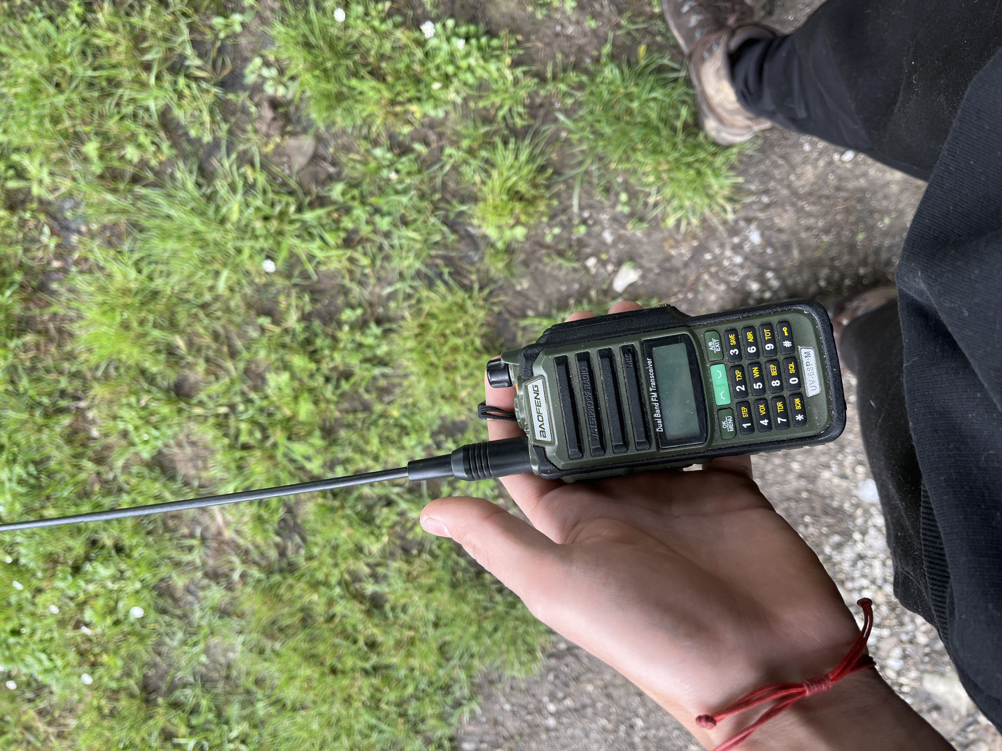 A person is holding a Baofeng brand, dual-band FM transceiver with an extended antenna in their hand. The device is dark green with black accents, has a screen, and a number keypad. The person wears a red string bracelet on their wrist and is dressed in black clothing. In the background, there is patchy green grass and a gravel path visible. There is no visible evidence to suggest this photo was taken in Shopite, Bulgaria.