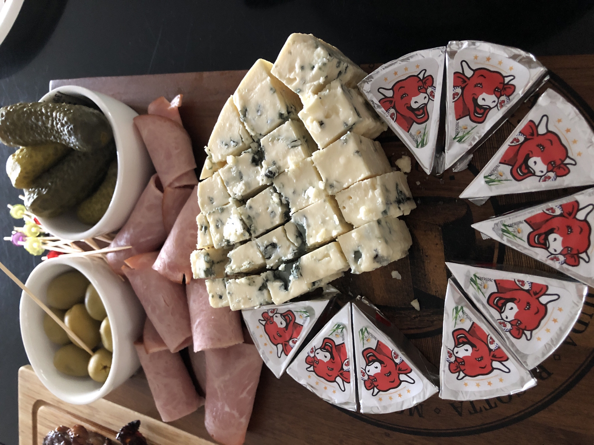 A high-angle shot captures a charcuterie board on a dark wooden surface, likely a table, possibly in a home or restaurant in Innisfil, Canada. The board is laden with various food items. In the upper left corner, a white ceramic bowl contains several green olives, with a wooden skewer protruding from it. To its right, another white bowl holds a pile of gherkin pickles, also pierced by a skewer. In front of these bowls, slices of pink ham are artfully rolled and arranged.
The central focus of the image is a generous portion of blue cheese, cut into numerous bite-sized cubes, revealing its distinct blue veins. Surrounding the blue cheese on the lower half of the board are several triangular wedges of cheese. These wedges are individually wrapped in foil featuring a cartoon illustration of a red cow. The foil wrappings are arranged in a circular fashion. The scene is well-lit, with natural light illuminating the food items, casting subtle shadows. The background is a dark, out-of-focus surface, suggesting the board rests on a table. No people are visible in the image.