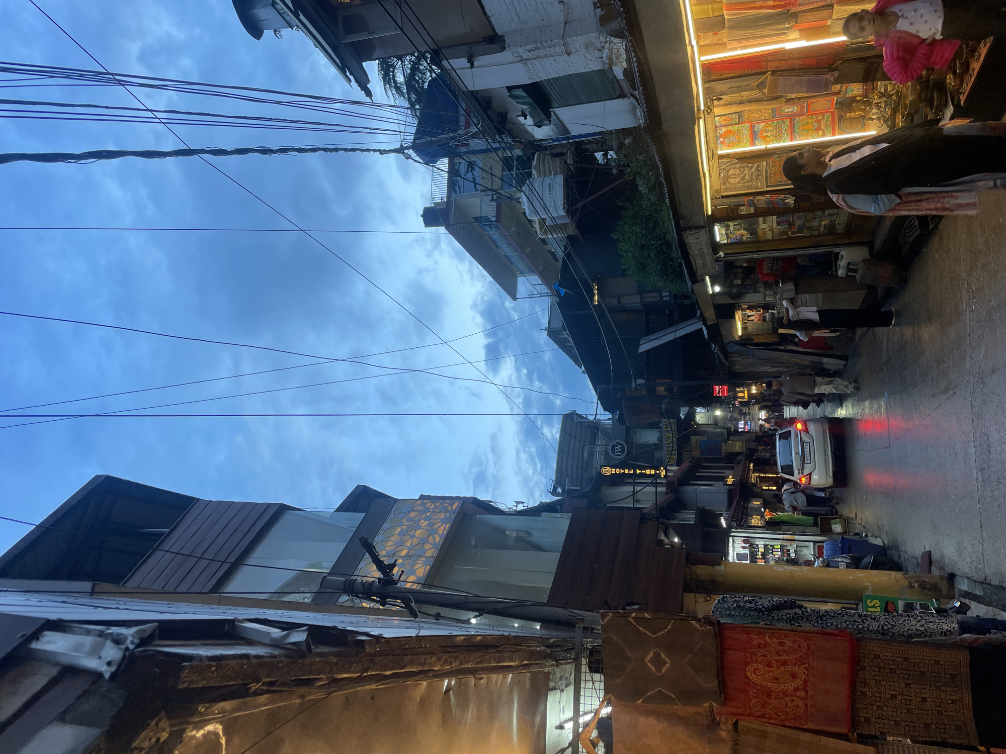 This image depicts a street scene in Bhagsu, India, characterized by narrow buildings lining a wet, reflective street. The time of day appears to be dusk or twilight, with a cloudy sky overhead and artificial lights illuminating storefronts.
On the left side of the street, the facades of multi-story buildings are visible, one with dark wooden paneling and large glass windows displaying intricate patterns. Another building features textiles hanging for display. The street itself is paved and glistens with moisture, reflecting the red taillights of a white car in the middle ground.
Several shops are visible, with illuminated displays showcasing various goods, including textiles, artwork, and what appear to be religious artifacts. People are present, some walking along the street, others interacting within or in front of the shops. In the foreground on the right, two young females are standing near a shop.
The environment is densely built with buildings clustered together, suggesting a commercial or residential area. Utility wires crisscross the sky, a common sight in many urban settings. The overall impression is one of a bustling, somewhat cluttered, but vibrant street scene in a hilly region.