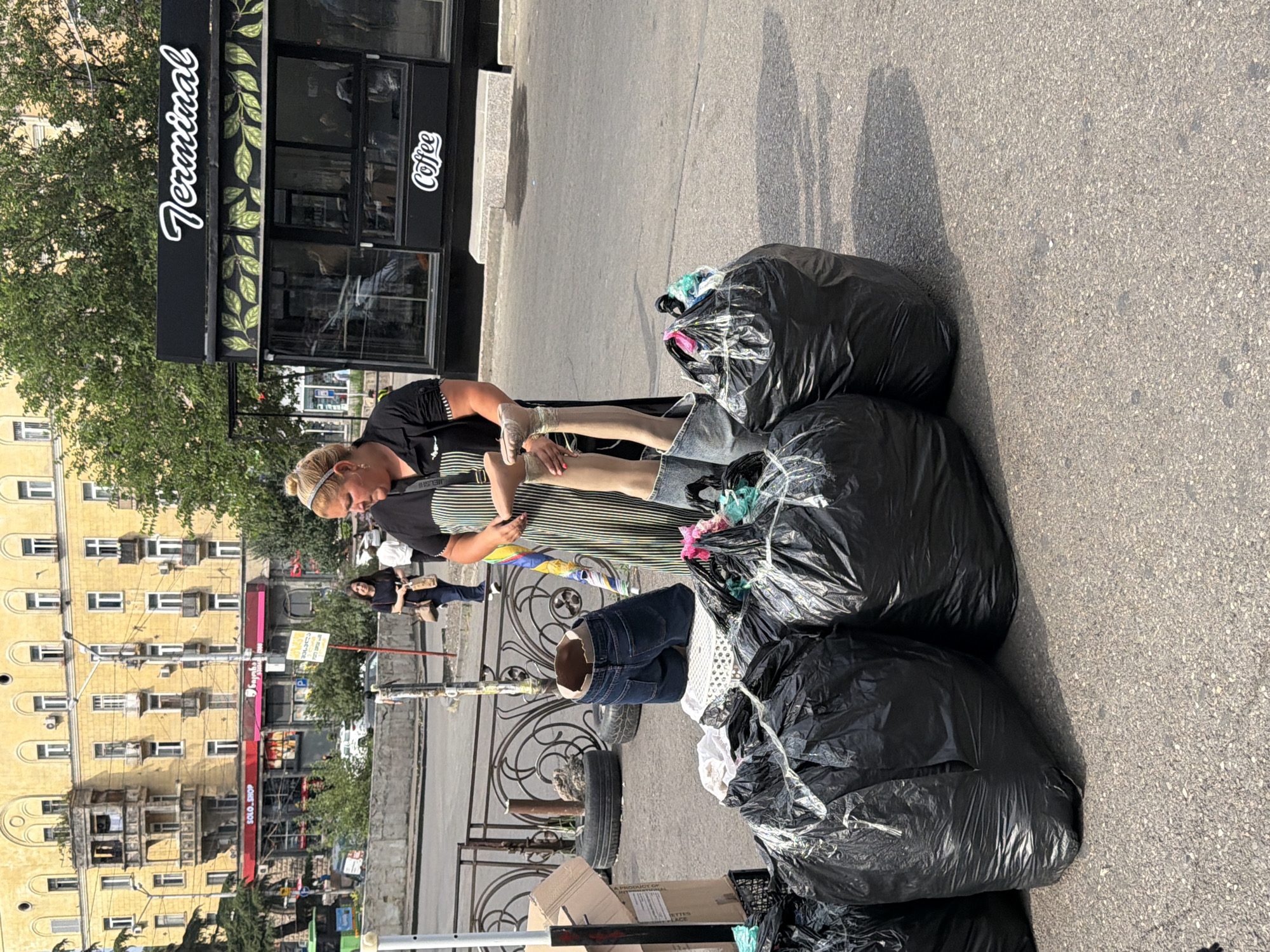In an outdoor setting on an asphalt surface, likely a sidewalk or street in Tbilisi, Georgia, a woman stands next to several large black trash bags. The woman is wearing a black and white striped apron over a black shirt, her blonde hair pulled back. She is holding up two mannequin legs, which are wearing denim shorts and striped socks. One leg is bent at the knee, with the foot pointing upwards. Several black trash bags are piled on the ground, some appearing to contain clothing or fabric. Behind the woman, a black kiosk with the word "Terminal" in white script and "Coffee" below it is visible. In the background, a yellow building with multiple windows and balconies suggests an urban environment, and trees are present. The overall scene appears to be a street vendor or market area.