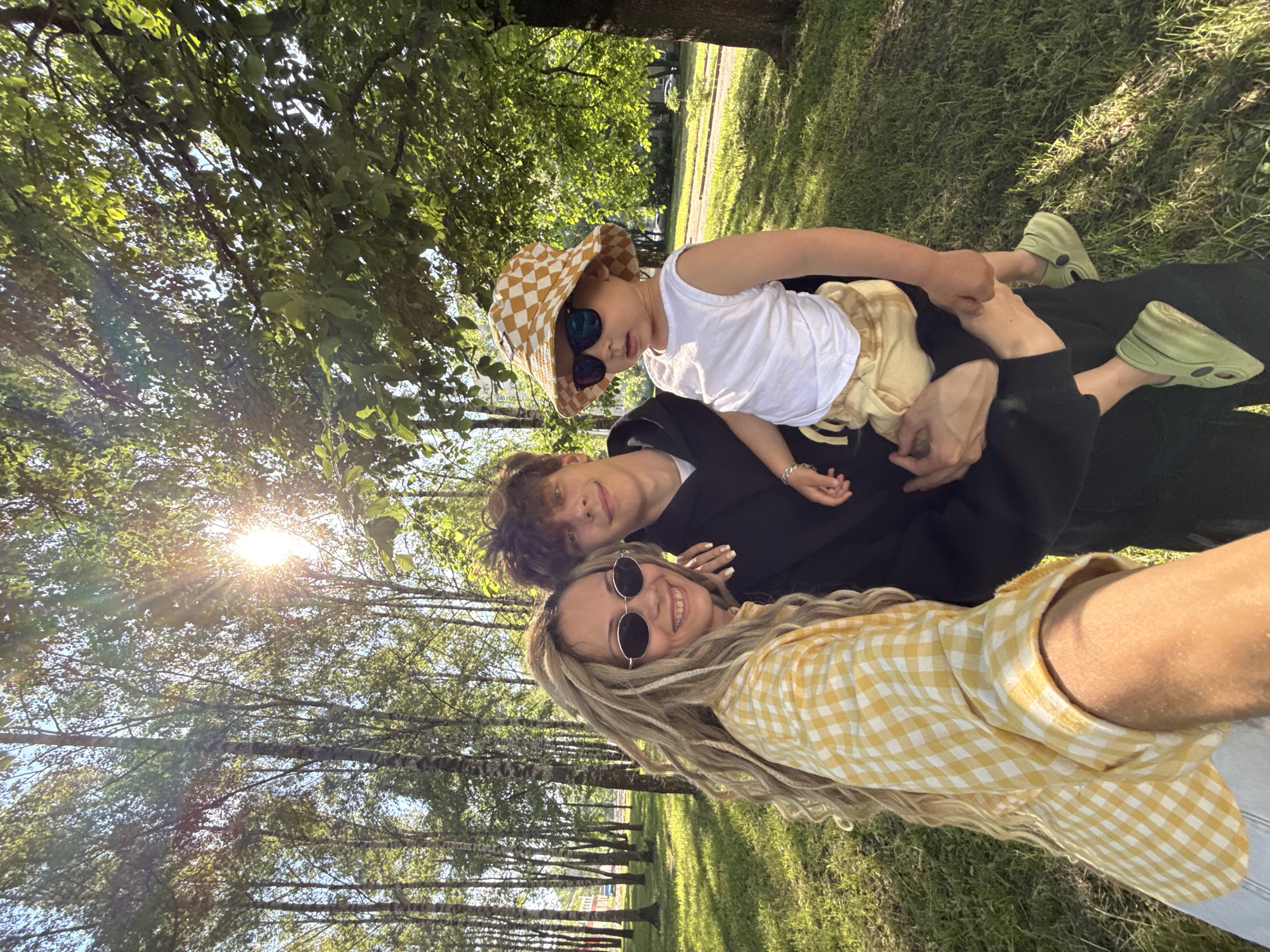 In an outdoor park setting, three individuals are captured in a selfie. A woman with blonde dreadlocks and oval sunglasses smiles at the camera, wearing a yellow and white checkered shirt. Beside her, a young man with dark, messy hair, wearing a black hoodie, holds a small child. The child, wearing a checkered bucket hat, sunglasses, a white tank top, and light-colored shorts, is held securely in the man's arms. They are all standing on a grassy area with a line of birch trees in the background, dappled with sunlight.