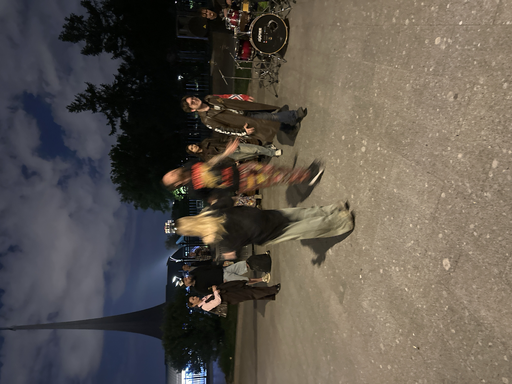 An outdoor scene captured at night or dusk shows a group of people gathered on a paved surface, with some appearing to be in motion. The image is oriented vertically, showing the ground on the right and the sky on the left.
In the upper central part of the image, a red drum set with a bass drum, snare, and cymbals is visible, suggesting a musical performance or rehearsal. The word "MAPEX" is clearly legible on the bass drum. Behind the drums, a person's head is partially visible. Standing closer to the viewer and to the left of the drums, a man with a beard and a long, dark brown coat is smiling towards the left. He wears jeans and dark boots. Further in front of him, another person in a dark shirt and jeans is blurred due to motion.
Towards the bottom right, a person with long blonde hair, wearing a dark top and light-colored pants, is also motion-blurred, suggesting movement. Closer to the foreground, a man with a beard, wearing a dark top, stands next to a person in a dark coat and a light pink shirt, who is holding a microphone. Other individuals are partially visible in the background, including one person in a light blue shirt and shorts.
The setting appears to be an urban outdoor space, possibly a plaza or park. Tall, dark trees frame the top and bottom left of the image. The sky is dark with visible clouds. In the far distance on the left, a tall, slender, dark structure or monument can be seen against the cloudy sky. The ground is a light gray, textured pavement. The overall ambiance suggests a casual public gathering or performance during the evening, possibly in Moscow, Russia.