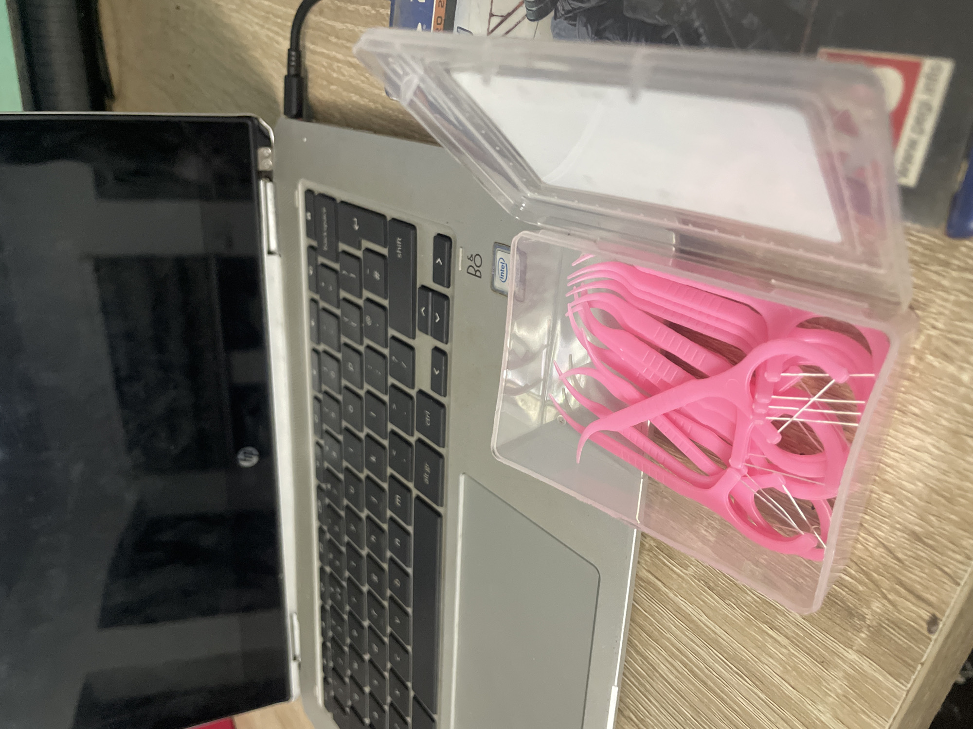 An overhead shot reveals a silver laptop and a clear plastic container filled with pink dental floss picks, arranged on a light-colored wooden desk. The laptop, possibly an HP model given a faint logo reflection on its black screen, has black keys with white lettering, including visible labels for "backspace," "shift," "ctrl," and "alt gr." Brandings "B&O" (likely Bang & Olufsen) and "intel" stickers are visible on the laptop's body. A black power cable is connected to the laptop's upper left side.
The open plastic container houses numerous bright pink dental floss picks, with their white floss strings distinctly visible. The desk surface features a light wood grain. In the background, some indistinct items, possibly books or other objects, are out of focus. The scene is static, suggesting an indoor setting like an office or home desk. No people are present, and there are no discernible cues regarding the time of day or specific emotions. The context provided indicates the location is Abuja, Nigeria.