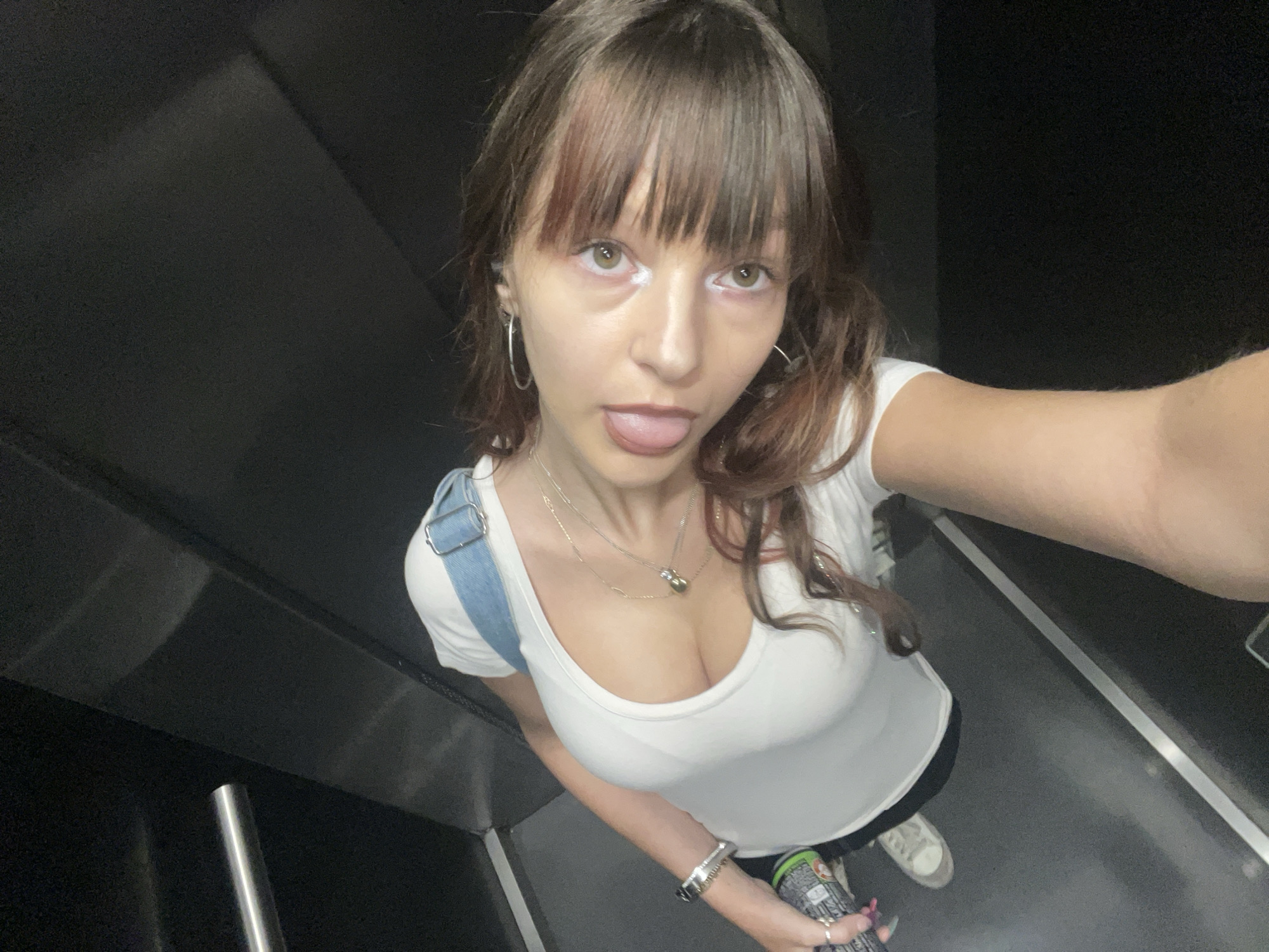 The image shows a young woman standing in what appears to be an elevator. She is wearing a white t-shirt, denim overalls, and sneakers. She has brown hair with bangs and is sticking out her tongue. She is holding a can in her right hand. The background is dark and industrial. There is no indication of the location being Raanana, Israel.