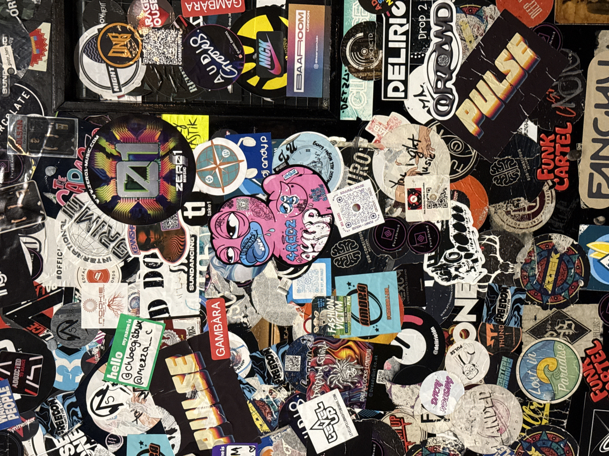 The image shows a dense collage of stickers on a dark surface, likely a wall or board. The stickers are of varying sizes, shapes, and designs, featuring logos, text, and graphics related to music, nightlife, and fashion brands. There are stickers representing specific DJs, events, and record labels, with words such as "FUNK," "HOUSE," "DISCO," "TECHNO," and "GROOVES" prominently visible, indicating the association with electronic music genres. "BAAROOM LONDON" is seen on one sticker, suggesting a location in the city of London, United Kingdom. Further emphasizing this locale are other stickers such as "Dreadlocks London". QR codes are also present on multiple stickers, potentially linking to online content. The stickers are layered and overlapping, creating a visually busy and vibrant composition.