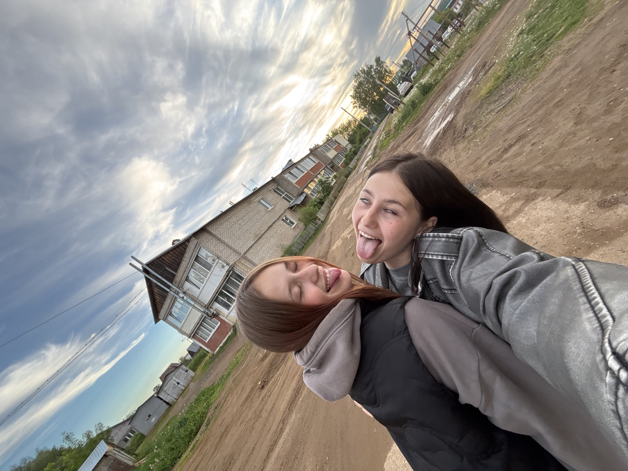 Two young women are taking a selfie outdoors. One is wearing a grey jacket and is sticking out her tongue with her eyes open. The other is wearing a grey hoodie and a black vest, and she is sticking out her tongue with her eyes closed. They appear to be standing on a dirt road in a rural setting. In the background, there is a two-story brick building, power lines, and some other houses. The sky is cloudy with a patch of sunlight. The photograph appears to have been taken in Фоки, Russia, given the request.
