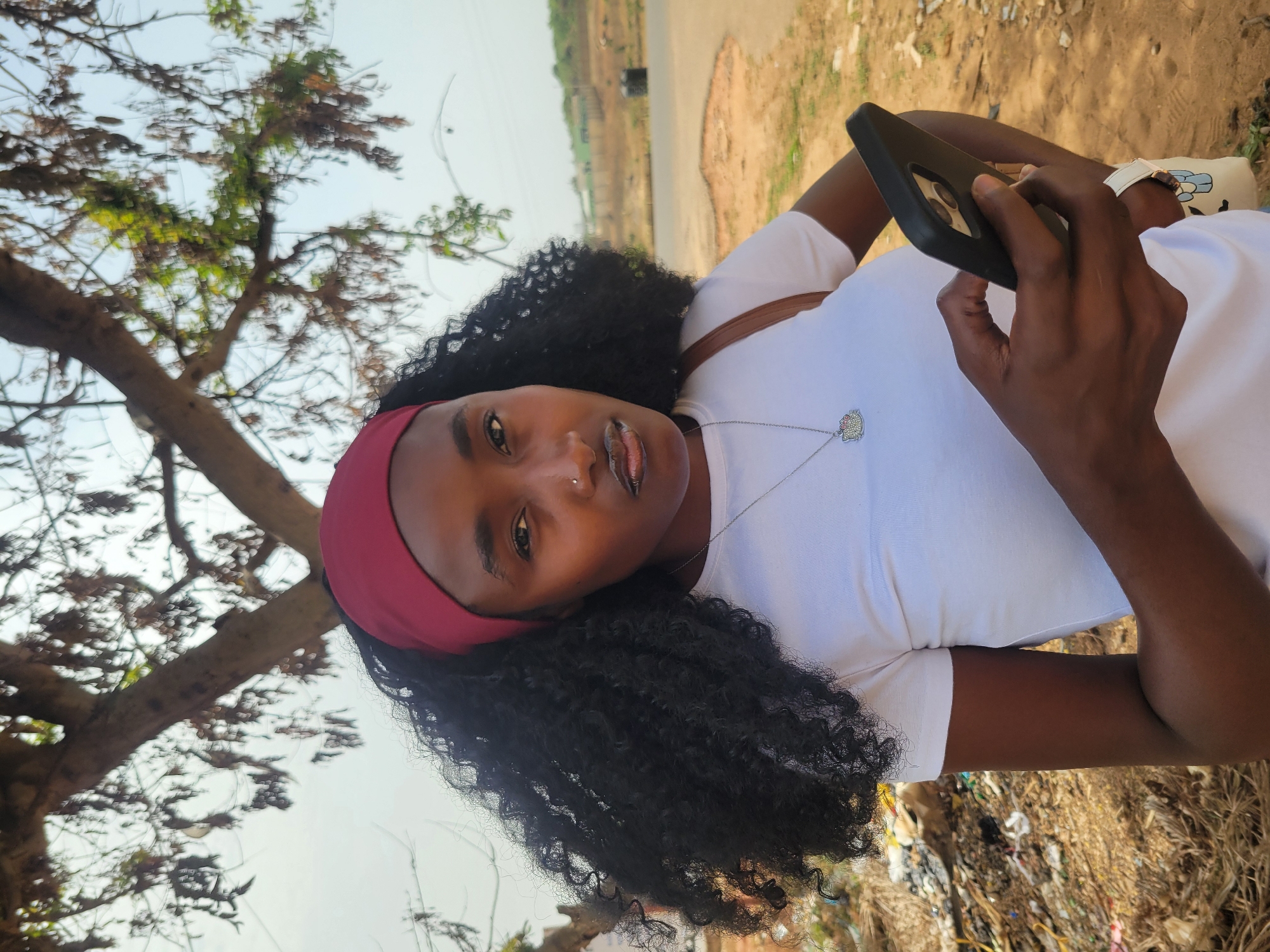 A medium shot shows a young Black woman with dark curly hair and a red headband looking at the camera, holding a black smartphone. She wears a white t-shirt and a silver necklace. The background reveals a tree on the left and a road leading into the distance. The location appears to be a grassy area with scattered debris. This photograph was taken near Jos, Nigeria.