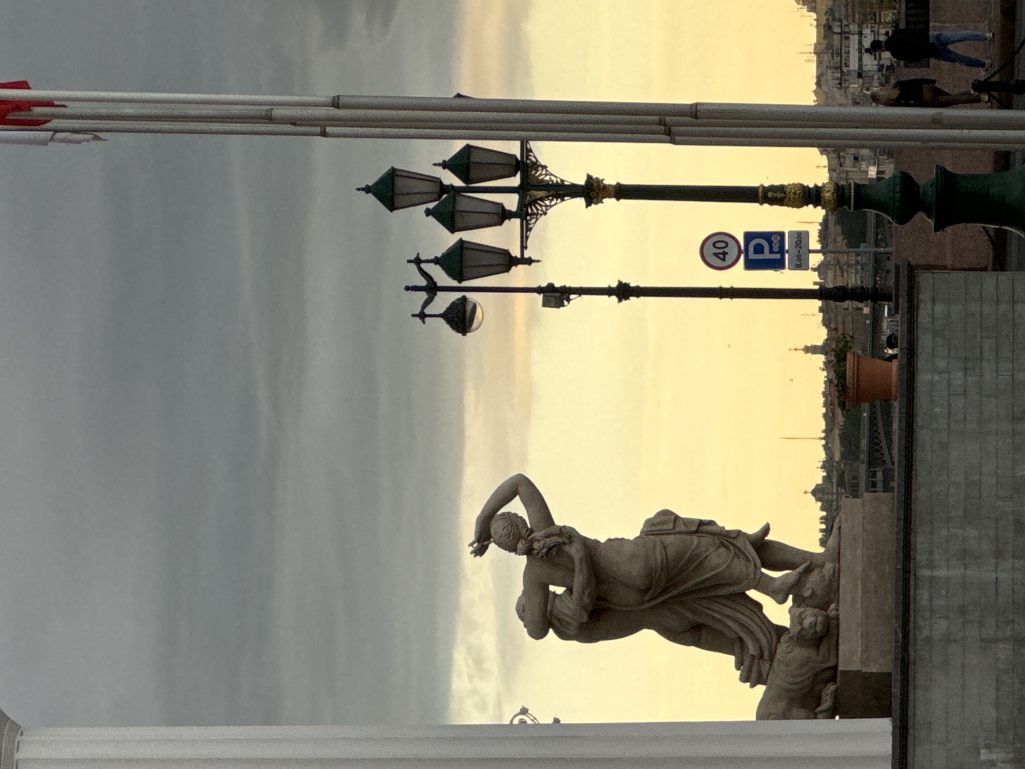 This is a medium shot, eye-level, vertical-format photograph of a cityscape at dusk. The sky is overcast with gray clouds and a pale yellow glow from the setting sun. In the foreground, on the left, is a large fluted white column. To its right is a classical stone sculpture of two figures, a man and a woman, embracing. The woman is reaching up to the man's head, while he holds her around the waist. They are standing on a stone pedestal, which rests on a gray stone wall. A stylized lion lies at the feet of the figures on the pedestal.
Behind the sculpture, to the right, are several ornate street lamps with green tops. The nearest lamp is a multi-headed one with four lights. Further in the background, a cityscape is visible with buildings and spires. A traffic sign with "40" in a red circle above a blue parking sign with "P" and times "8.00-20.00" is positioned between two street lamps.
In the far right of the image, two individuals, a woman with blonde hair and a man in a dark jacket and jeans, are walking away from the camera. Another individual in a dark jacket and jeans is walking towards the camera. The photograph captures the atmosphere of Saint Petersburg, Russia, during twilight hours, showcasing a blend of historical monuments and urban life.