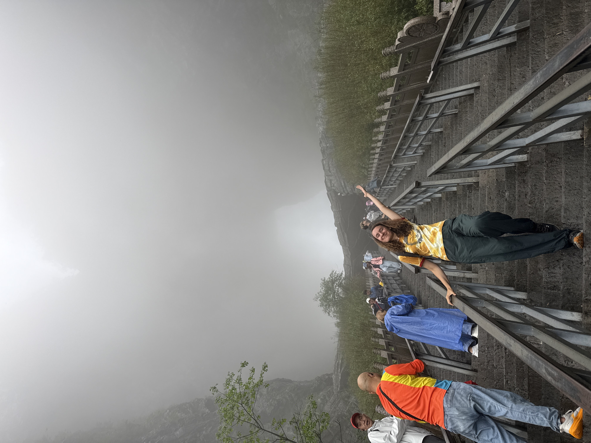 The image shows a group of people on a stone staircase with metal railings, descending into dense fog. The location is likely in Zhangjiajie, China, given the mountainous terrain and characteristic fog.
A woman in a yellow patterned shirt and dark pants is posing with her arm raised. Below her, others are walking down the steps, including a person in a blue raincoat. At the bottom of the frame, a man in an orange and yellow shirt is partially visible, along with another person in a white shirt and a red hat.
The environment is characterized by heavy fog obscuring the landscape. Vegetation is visible on the sides of the staircase, adding to the natural setting.