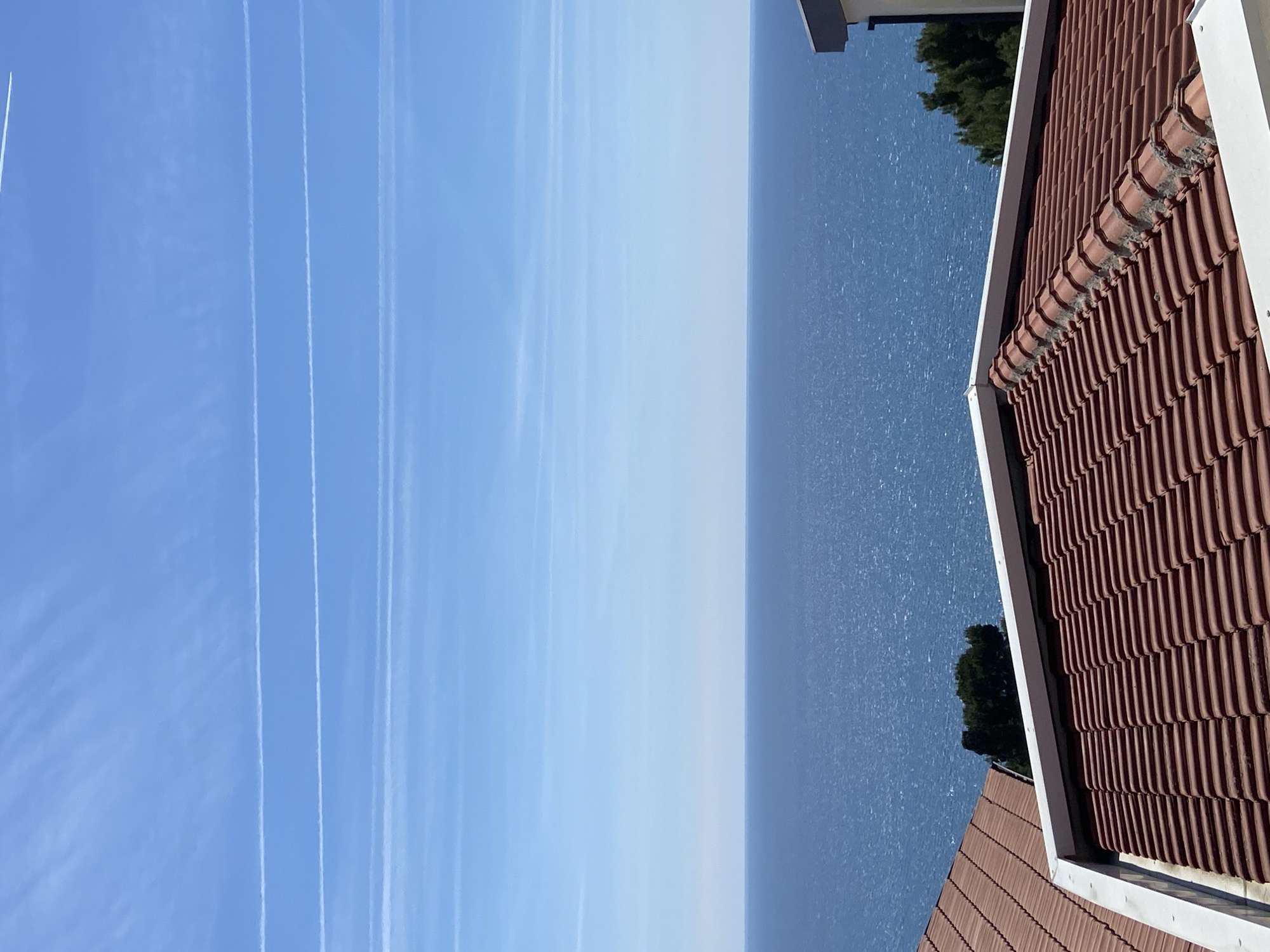 The image shows a view from a building in Šušanj, Montenegro. The scene captures the Adriatic Sea under a blue sky marked with contrails from airplanes. In the foreground, a portion of a terracotta tile roof and a white gutter are visible. Beyond the roof, the blue water of the sea stretches to the horizon. Patches of dark green vegetation are situated in the space between the roof and the sea. The sky is bright blue and largely clear with several contrails streaking across it.