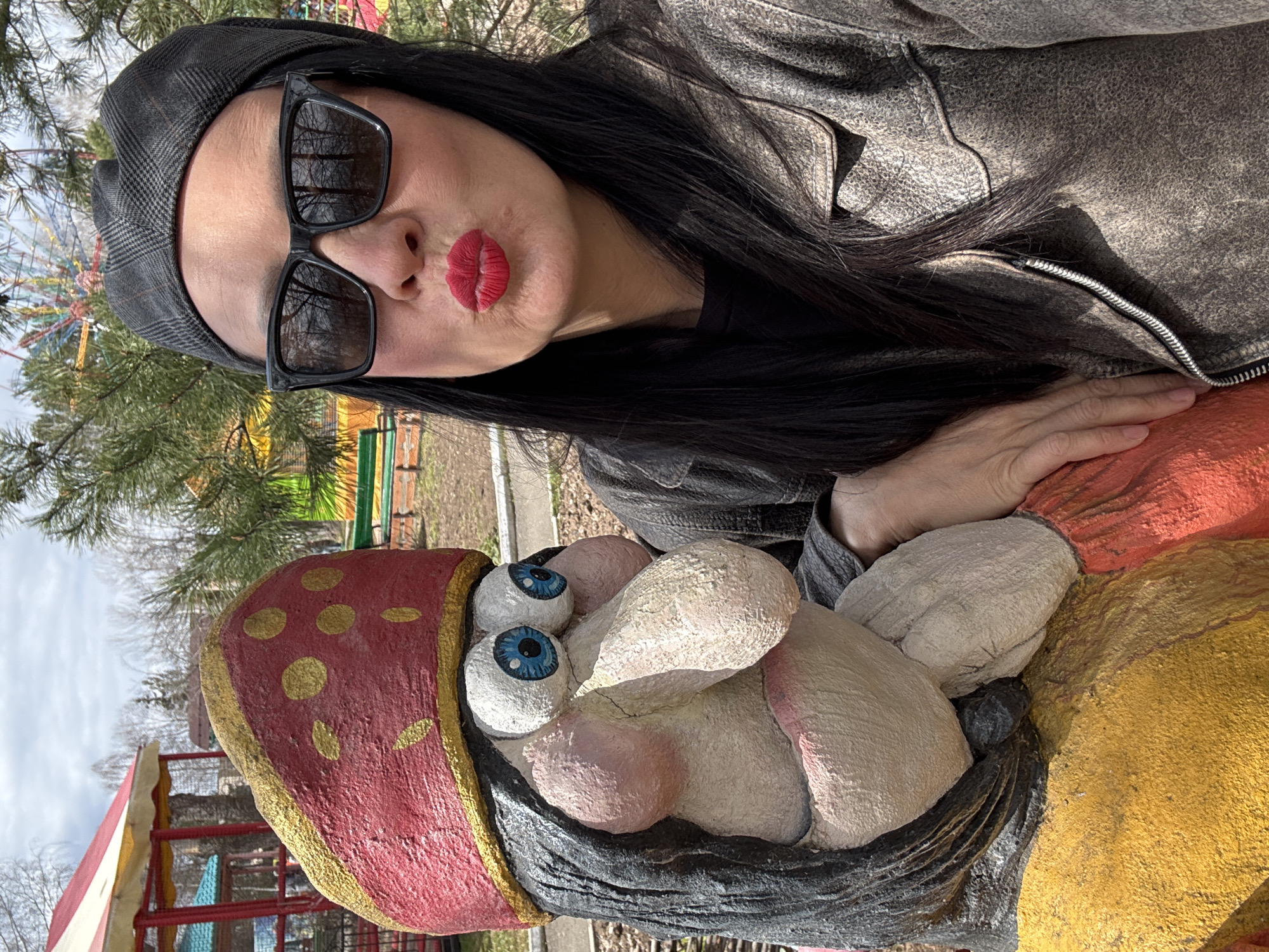 A woman with long, dark hair, wearing a grey hat and sunglasses, poses with pursed red lips in front of a painted cement sculpture of a gnome or dwarf with large, bulging eyes. The sculpture has a red hat with gold polka dots, a black beard, and is painted with yellow and orange colors. The photo appears to be taken in a park in Chaikovsky, Russia, based on the style of the sculpture, which is typical of the area. A Ferris wheel and colorful park structures are visible in the background. The woman's hand is resting on the sculpture. She is wearing a grey jacket. The sky is overcast.