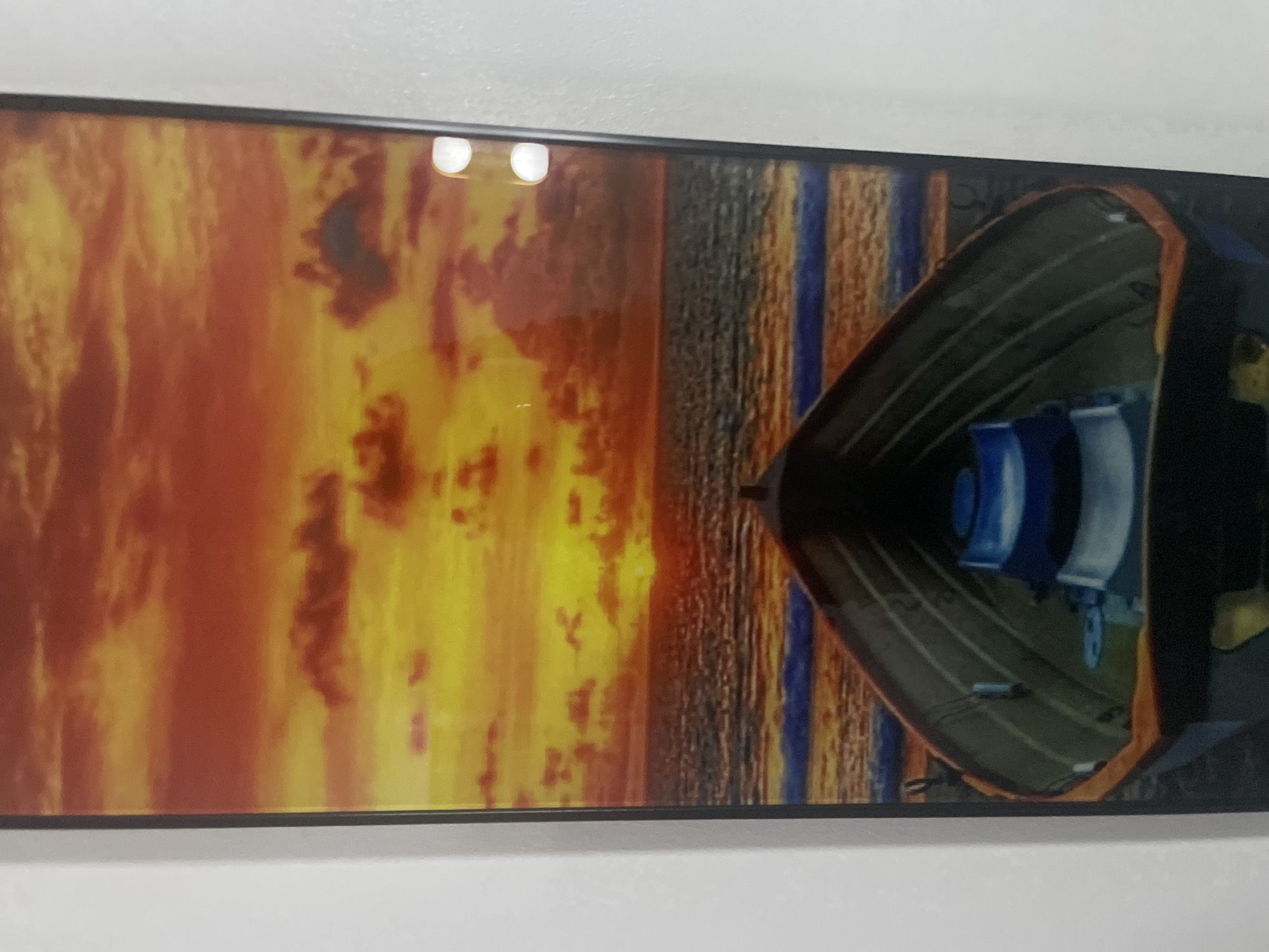 The image shows a framed print or painting. The artwork depicts a boat interior facing the viewer, with a sunset or sunrise scene in the background. The background shows an orange and yellow sky with textured brush strokes, possibly representing clouds. Vertical blue and brown stripes are also visible, suggestive of a body of water reflecting the sky. The boat is painted with green and orange sides. Two cylindrical objects painted blue and white are placed within the boat. The frame surrounding the print is black. The print is mounted on a white wall. Location is not discernible.