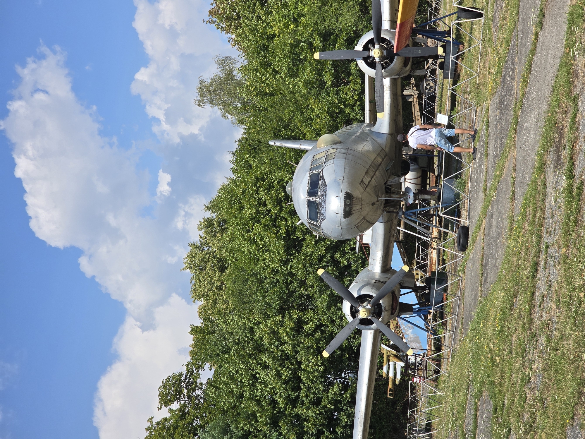 In Pustiměř, Czechia, an outdoor museum setting displays a vintage twin-engine airplane, likely a relic from a previous era, situated amidst lush green trees under a partly cloudy sky. The aircraft, with its silver fuselage and propellers, is elevated on support structures, suggesting it is part of an exhibition. A lone male figure, wearing a white t-shirt and blue shorts, stands facing the aircraft, his back to the camera, observing the display. A low metal barrier separates the public from the exhibit. The ground is a mix of concrete and grass, with scattered wildflowers adding a touch of nature to the scene. The overall atmosphere appears tranquil and educational, inviting viewers to explore aviation history.