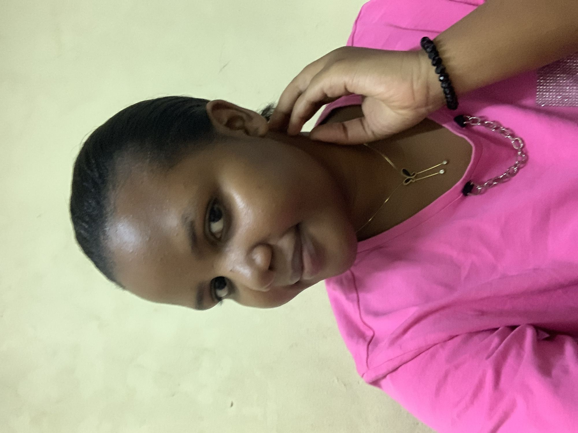 A woman is posing for a picture. She has dark hair, a light brown complexion, and is wearing a bright pink shirt. She is also wearing a gold necklace with a pendant that looks like a pair of scissors, and a black beaded bracelet. Her hand is near her ear. The background is a plain, light yellow or cream color. There is no information to suggest that this picture was taken in Dar es-Salaam, Tanzania.