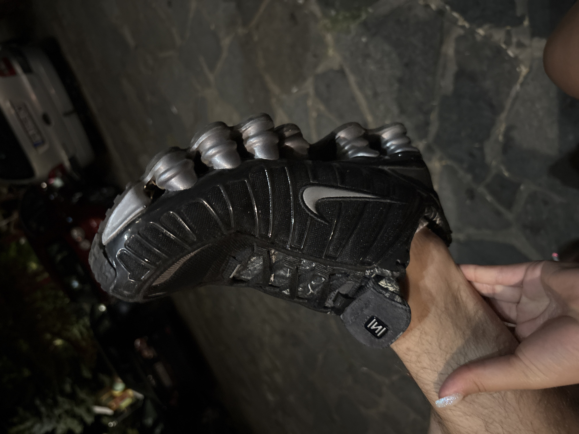 A black Nike Shox sneaker is held up against a background of parked cars and cobblestone ground, illuminated by artificial light. The shoe is worn and appears to be on a person's foot, with a hand visible in the foreground supporting it. The background shows a silver car and a darker car. The image is taken at night. The location is not specified to be Sopot, Bulgaria.
