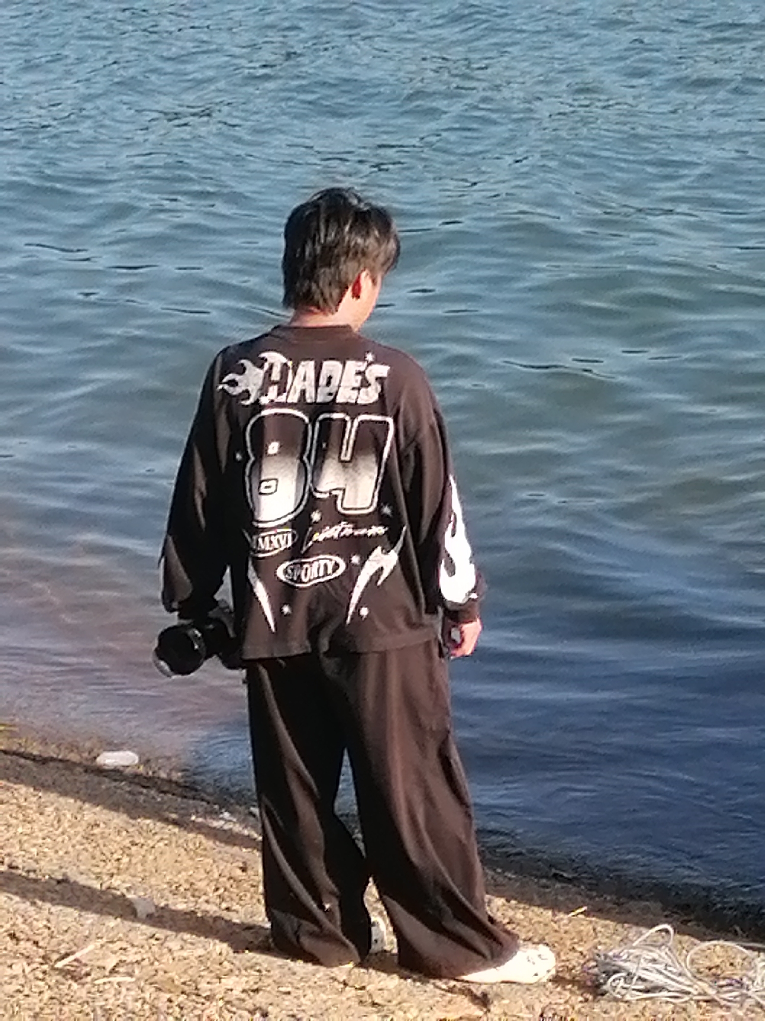 A person stands on the shore of Xuan Huong Lake in Da Lat, Vietnam, facing away from the camera. The person is wearing a black long-sleeved shirt with white graphic text and flames, and loose-fitting black pants. They are holding a black cylindrical object in their right hand. The ground is covered in small stones and sand, and a coiled rope is visible to the right. The water of the lake is calm with gentle ripples.