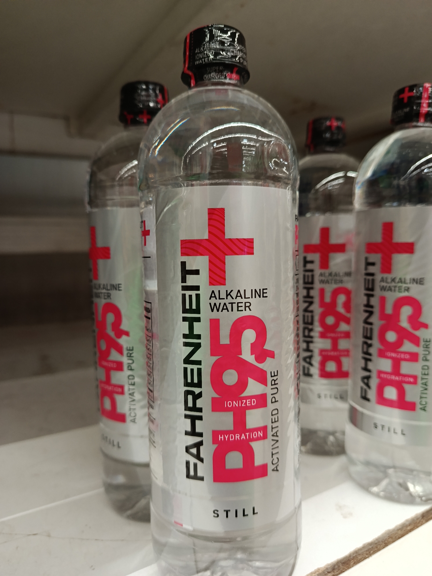 The image shows several bottles of "Fahrenheit PH95+" alkaline water on a shelf. The bottles have a silver label with a red plus sign and the brand name "Fahrenheit" printed vertically. The words "Alkaline Water", "Ionized Hydration", and "Activated Pure" are also prominently displayed in red on the label. The word "Still" is printed in a smaller font at the bottom of the label. The bottles have black caps with a red plus sign on top. The shelf appears to be in a store, but the location (Moscow, Russia) is not evident from the image.