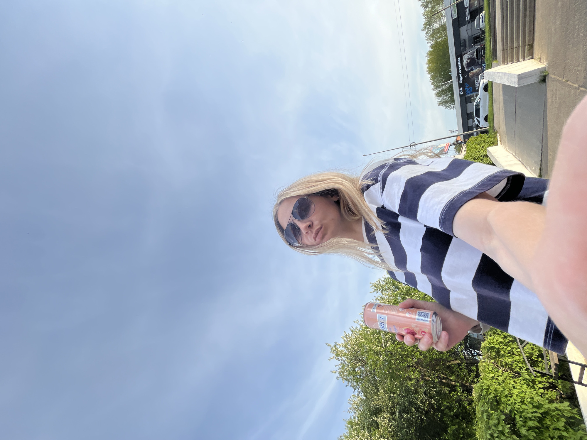 A young woman stands outside in Chaikovsky, Russia, holding a pink can. She wears sunglasses and a navy and white striped shirt. She is angled upward with the blue sky behind her. In the background is a two-story building with a sign in Russian, some trees, and a white car.
