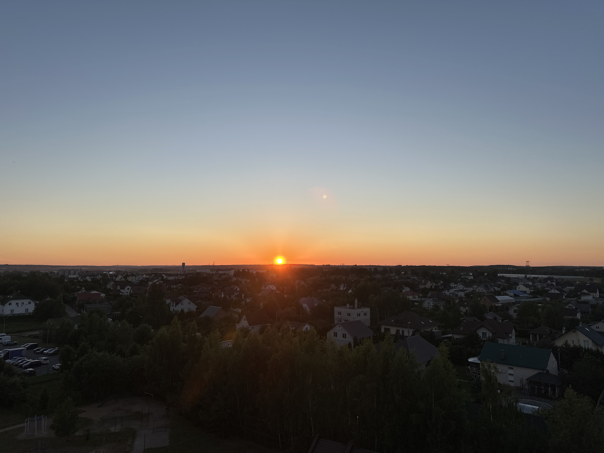 This aerial photograph captures a suburban landscape at sunset, likely in the vicinity of Шчомысліцкі сельскі Савет, Belarus. The sky transitions from a deep blue at the top to a warm orange hue near the horizon, where the sun, a small, bright disc, is setting. Below the sky, a sprawling residential area unfolds, characterized by numerous houses with varied rooflines and architectural styles, interspersed with dense clusters of trees. In the foreground, a darker area suggests a park or recreational space with some playground equipment. To the left, a parking lot is visible, filled with several parked cars. The overall scene depicts a tranquil evening in a settled, suburban environment under a dramatic sky.